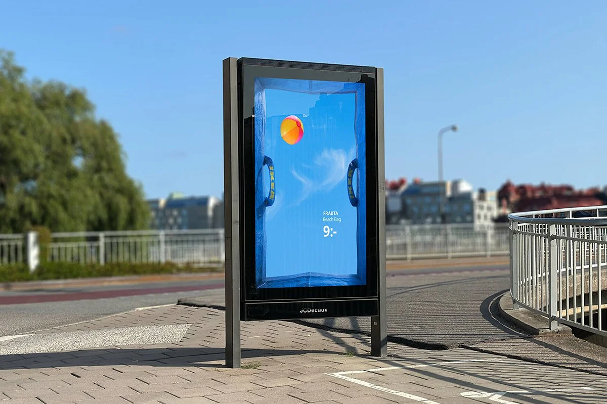  JCDecaux street billboard displaying IKEA FRAKTA campaign creative, featuring a blue bag design with beach ball visual, set on a sunlit urban street with railings and city buildings in the background. 