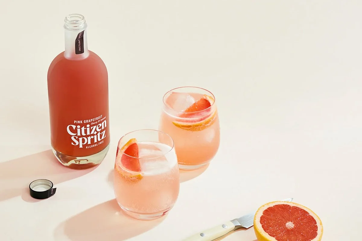  Citizen Spritz pink grapefruit alcohol‑free aperitivo bottle with two spritz cocktails garnished with citrus slices, styled on a clean neutral background for a fresh, low‑ABV drinking occasion. 