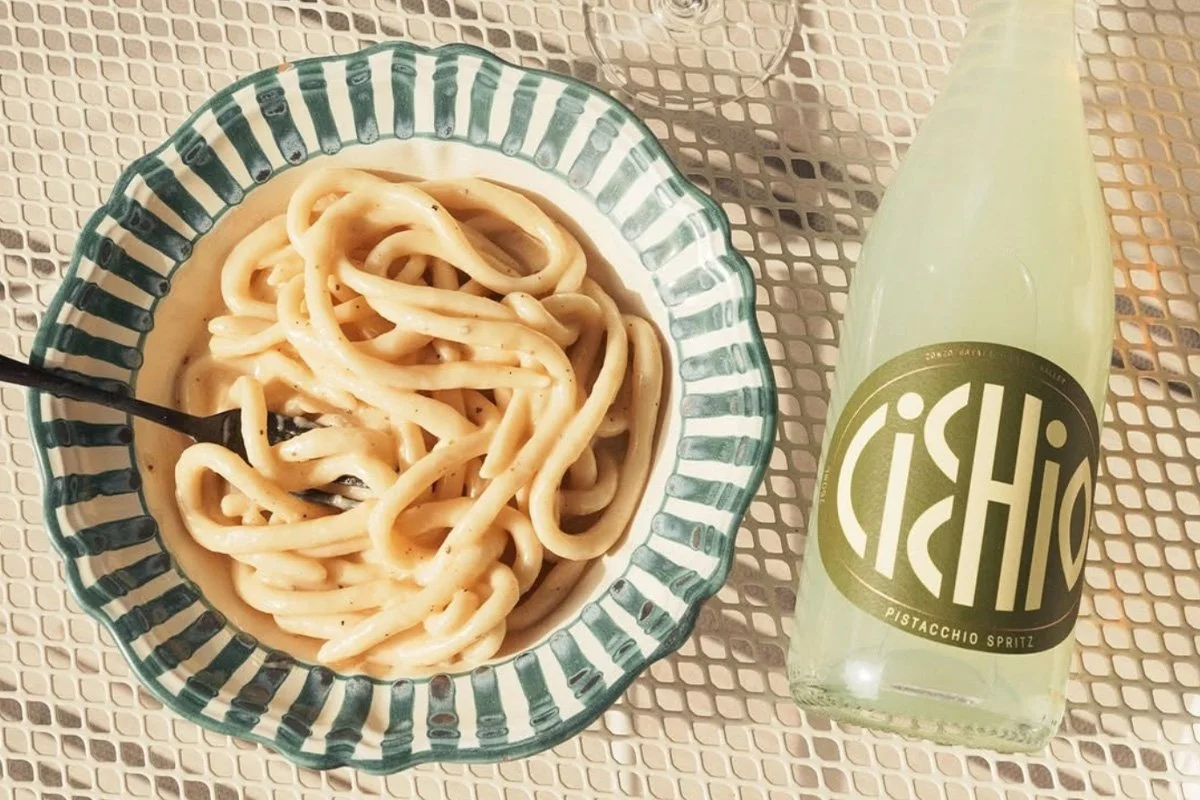  Bowl of creamy pasta served alongside a bottle of Cicchio pistachio spritz, a pistachio‑forward aperitivo, styled on a textured outdoor table to showcase a relaxed, modern aperitivo dining moment 