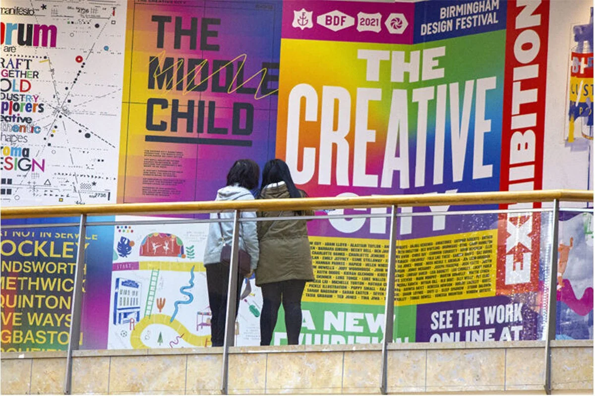  Large‑scale typographic posters displayed in a public space, featuring bold, colourful designs for Birmingham Design Festival and contemporary graphic design themes, viewed by visitors from a balcony. 