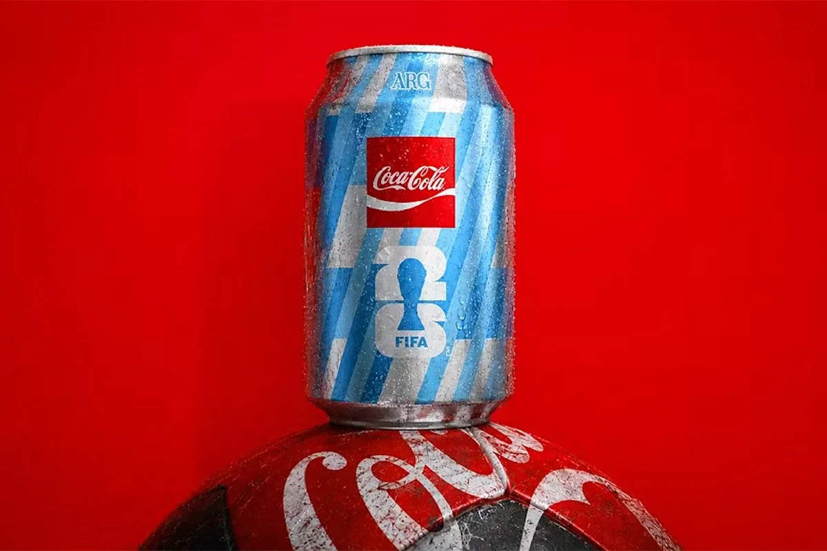  Limited edition Coca‑Cola FIFA can in blue design placed on a football against a bold red background, featuring condensation detail and sports branding tied to international tournament campaigns. 