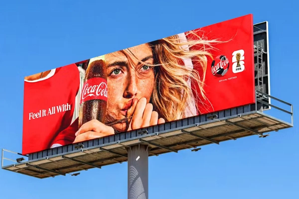  Coca‑Cola FIFA campaign billboard featuring a person holding a glass bottle against a bold red backdrop, with ‘Feel It All With’ messaging and official tournament branding displayed on a large outdoor structure. 