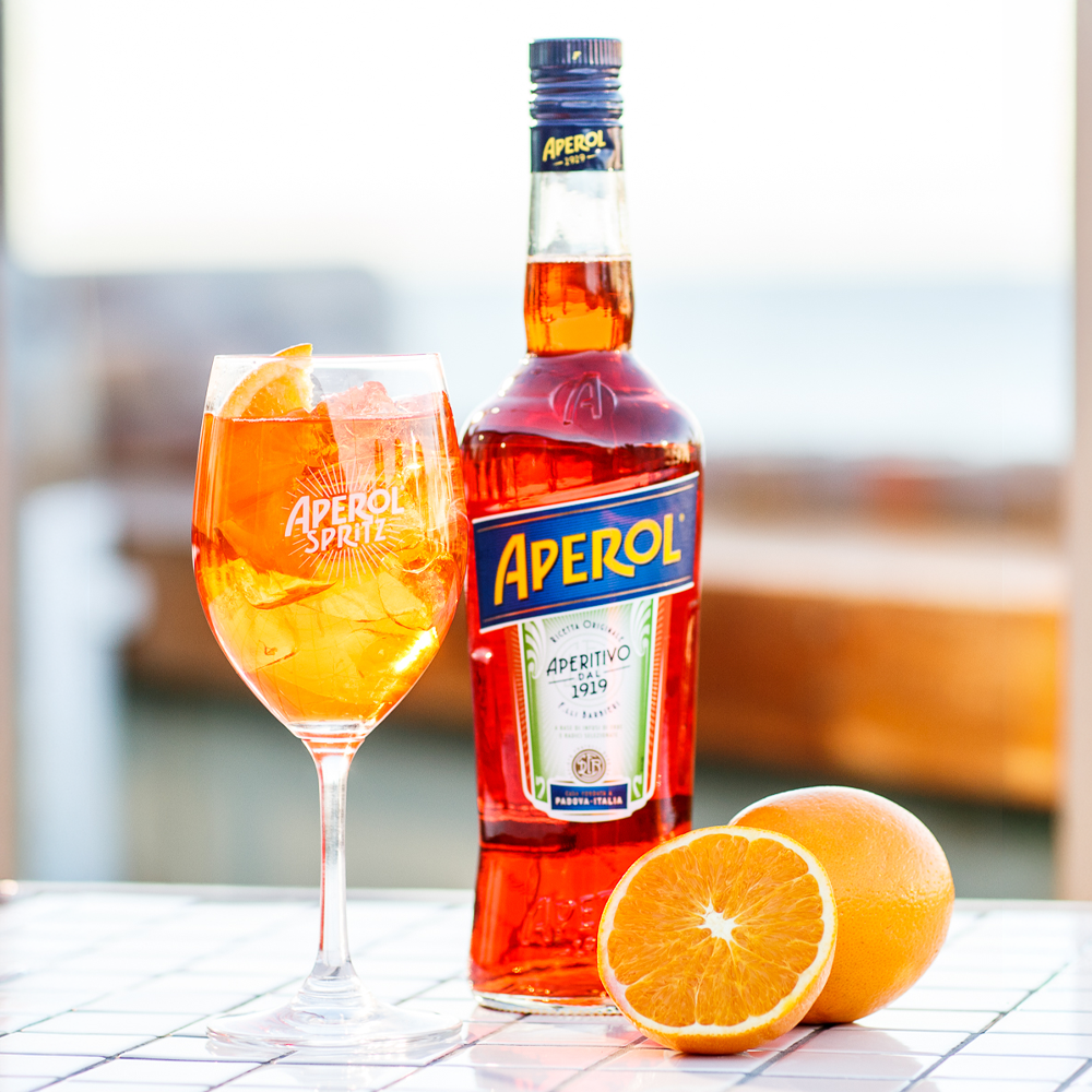  Aperol Spritz cocktail served over ice with an orange garnish beside a bottle of Aperol aperitif, showcasing the iconic Italian spritz serve in a bright outdoor setting. 