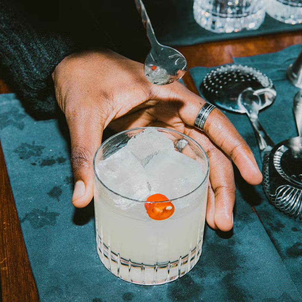  Soho House Picante cocktail served over ice in a cut-glass tumbler, garnished with a red chilli slice, as a bartender prepares the drink with professional bar tools in a premium hospitality setting. 