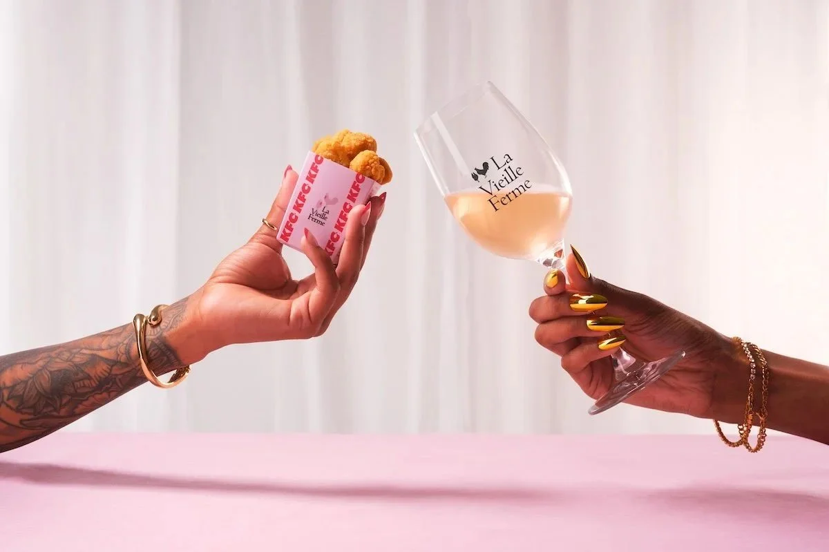  Hand holding KFC popcorn chicken beside a glass of La Vieille Ferme rosé wine, highlighting a food and wine pairing collaboration. 