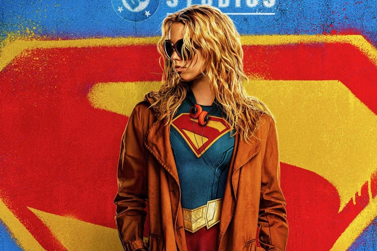 Promotional artwork for the Supergirl film featuring a character in a superhero costume and coat standing before the iconic Supergirl shield logo, using bold red, blue and yellow colours inspired by comic book design.