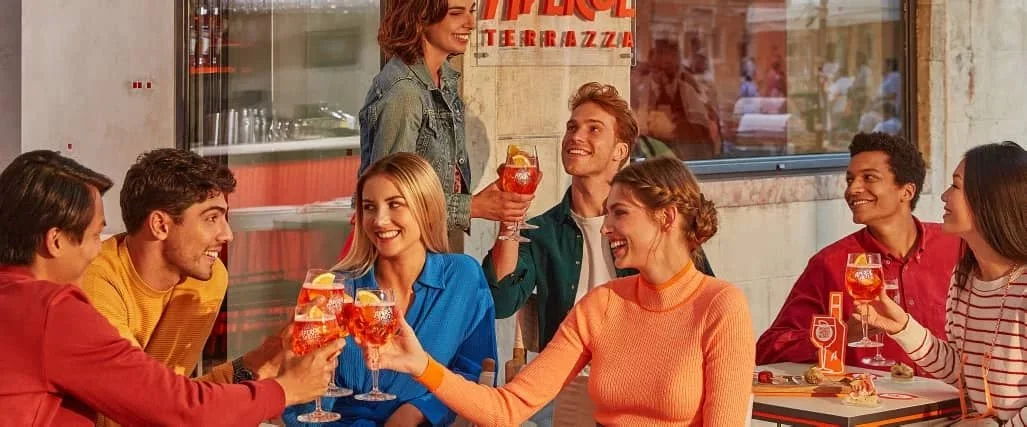 Group of friends gathered at a terrace bar, raising Aperol Spritz cocktails in branded glasses, with colourful outfits, shared plates and outdoor seating capturing a lively social aperitivo occasion.”