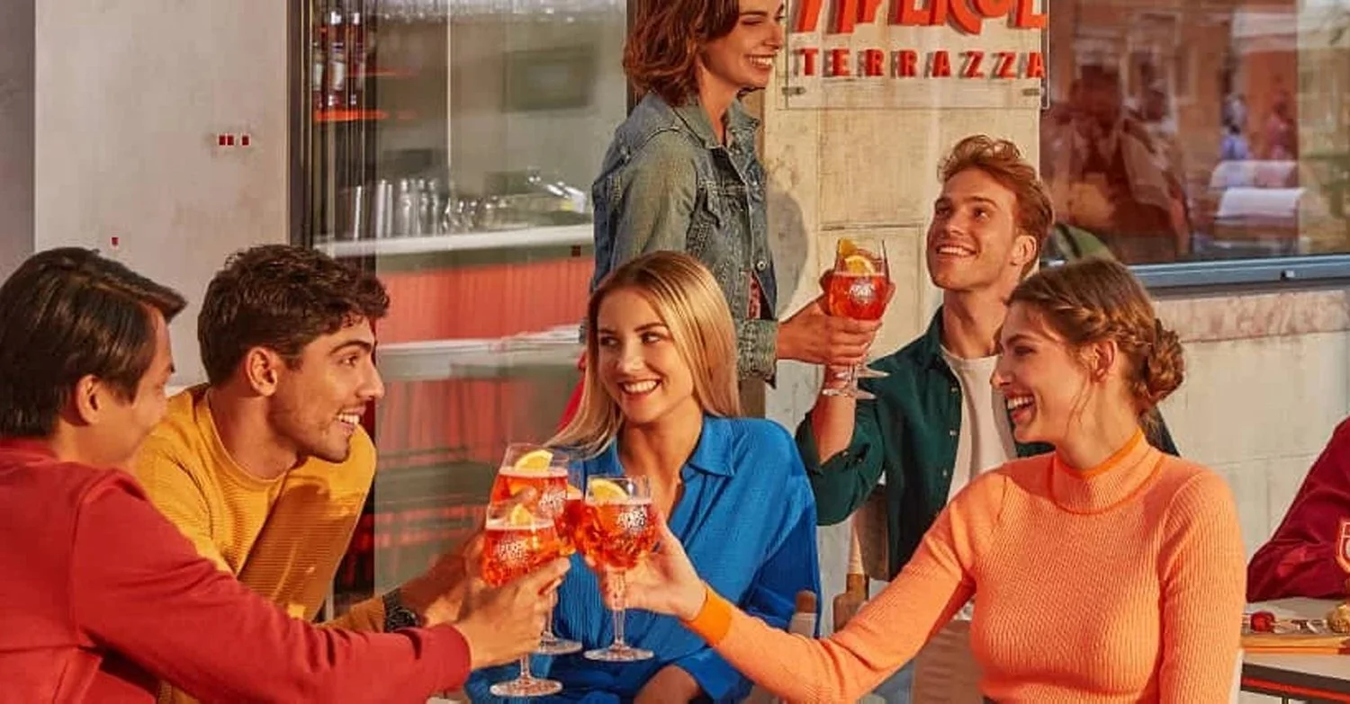 Group of friends gathered at a terrace bar, raising Aperol Spritz cocktails in branded glasses, with colourful outfits, shared plates and outdoor seating capturing a lively social aperitivo occasion.
