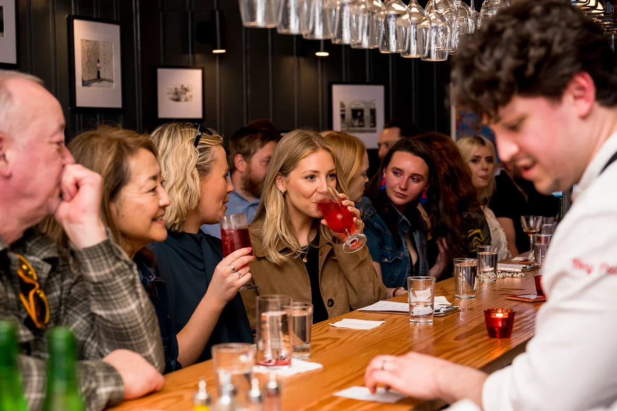 Guests seated at a busy bar during Edinburgh Bar Show 2026, sampling cocktails as a bartender serves drinks across a wooden counter, with glassware overhead and a lively industry event atmosphere.