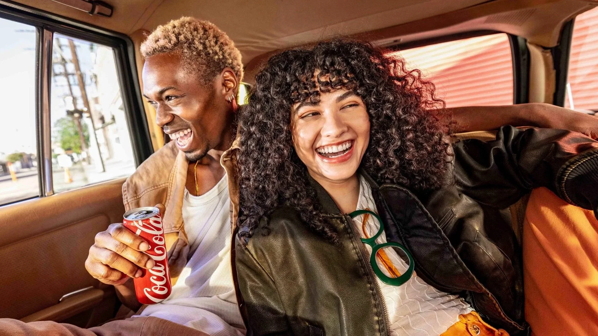  Two people seated in a car, with one holding a Coca‑Cola can, capturing a relaxed social moment and showcasing iconic red packaging in a lifestyle‑focused setting with warm natural lighting. 