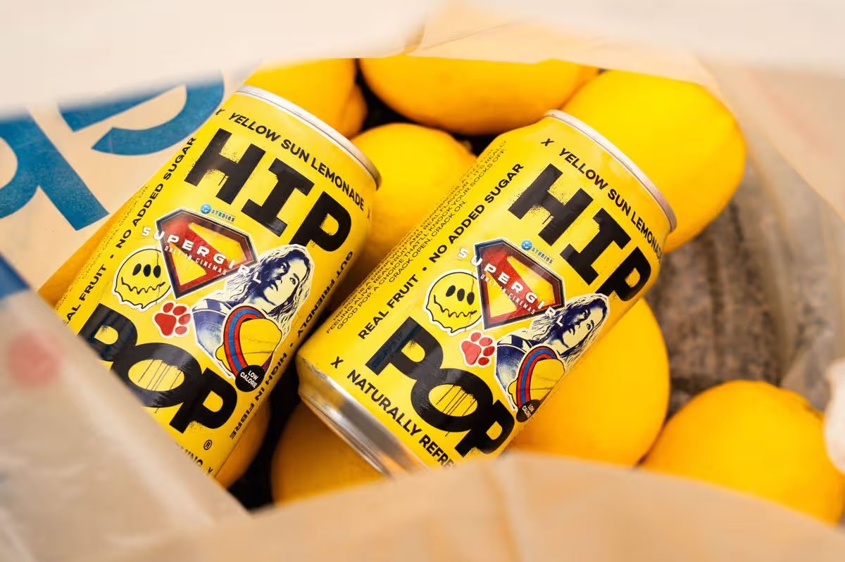 HIP POP Yellow Sun Lemonade cans from the limited‑edition Supergirl collaboration, displayed among fresh lemons to highlight real fruit ingredients, no added sugar credentials and vibrant comic‑inspired packaging design.