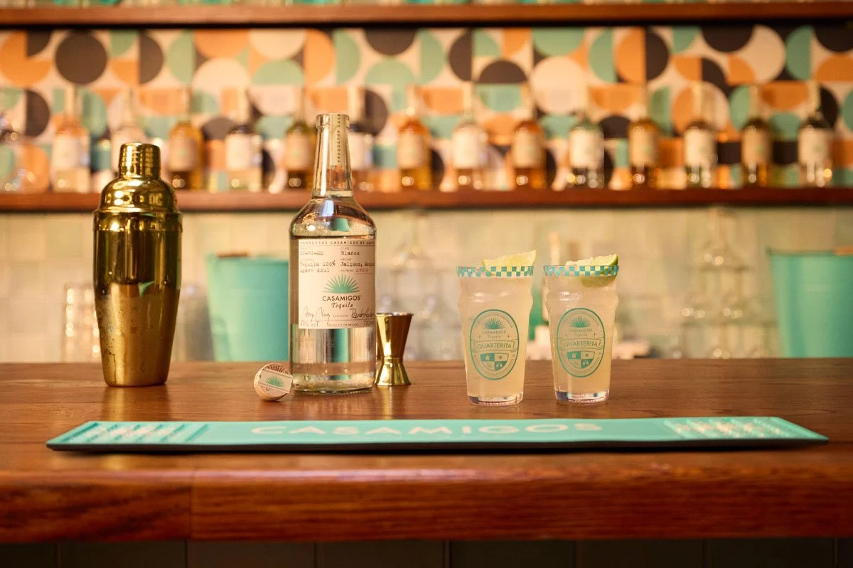 Casamigos Tequila bottle displayed behind a bar alongside two branded Quarterita cocktails, a cocktail shaker and bar tools, showcasing a premium tequila serve in a stylish hospitality setting.