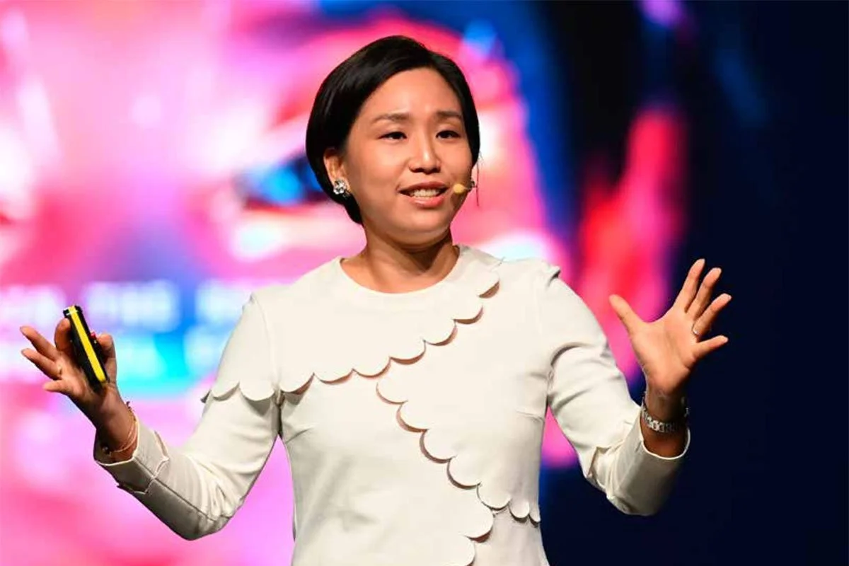 Hyunjin Kim, Professor of Strategy at INSEAD, delivering a presentation on stage at a global conference, holding a remote clicker against a vivid, colourful screen backdrop in a large auditorium setting.