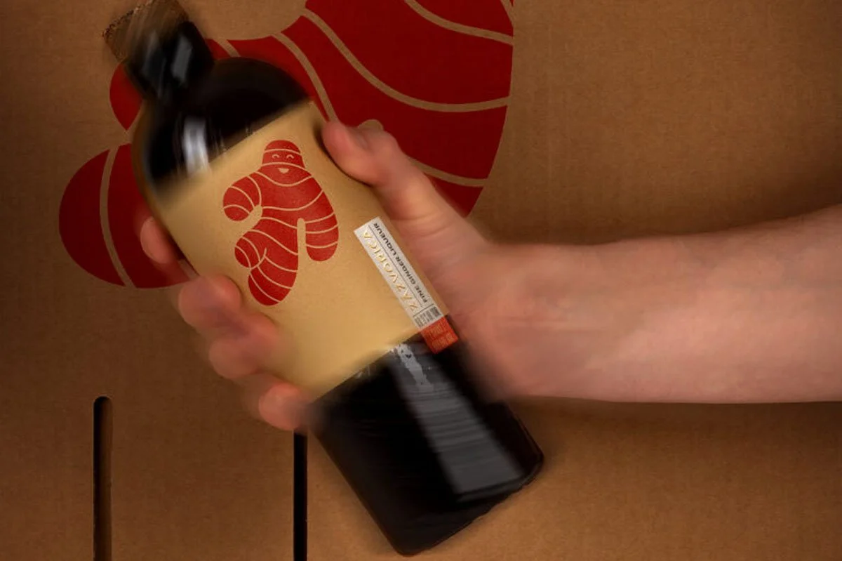 Hand holding and shaking a bottle of Zázvorica fine ginger liqueur against branded packaging, highlighting the distinctive ginger-inspired label design and the dynamic character of this traditional Slovak spirit.