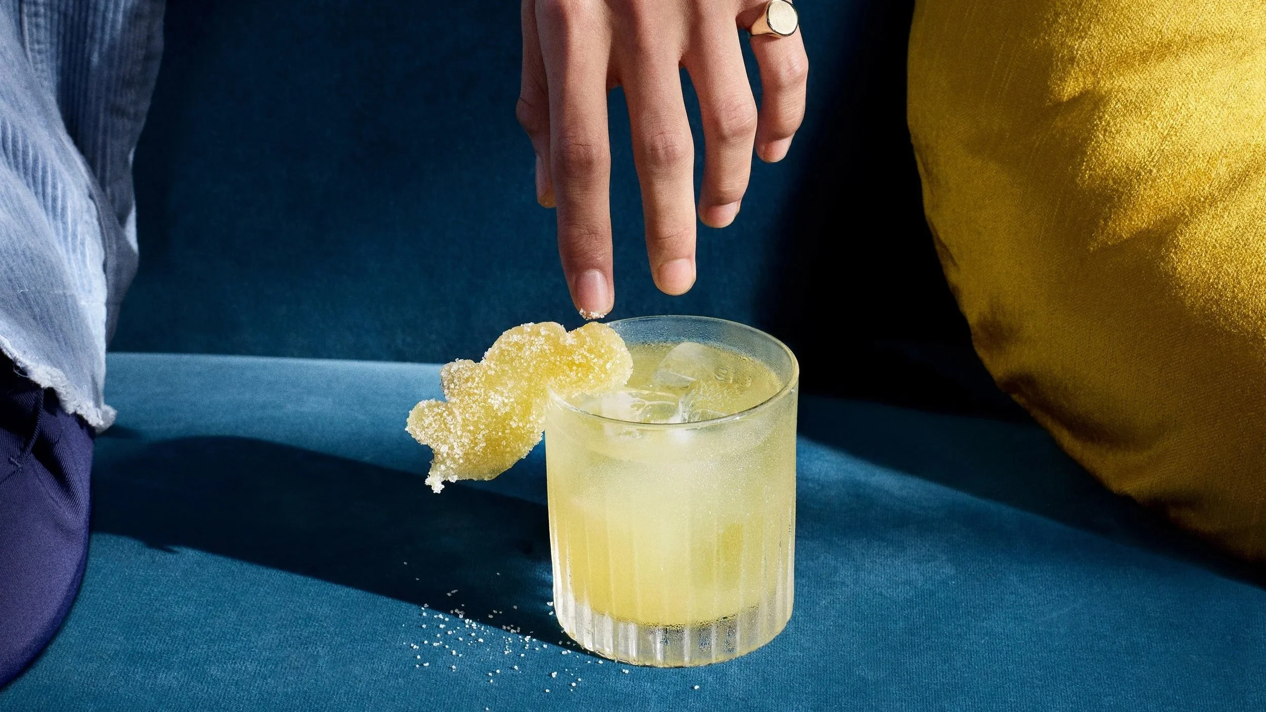 Pale yellow cocktail served over ice in a textured glass, garnished with a sugar‑coated citrus sweet, styled on a blue velvet sofa to showcase contemporary cocktail presentation and premium drinks culture.