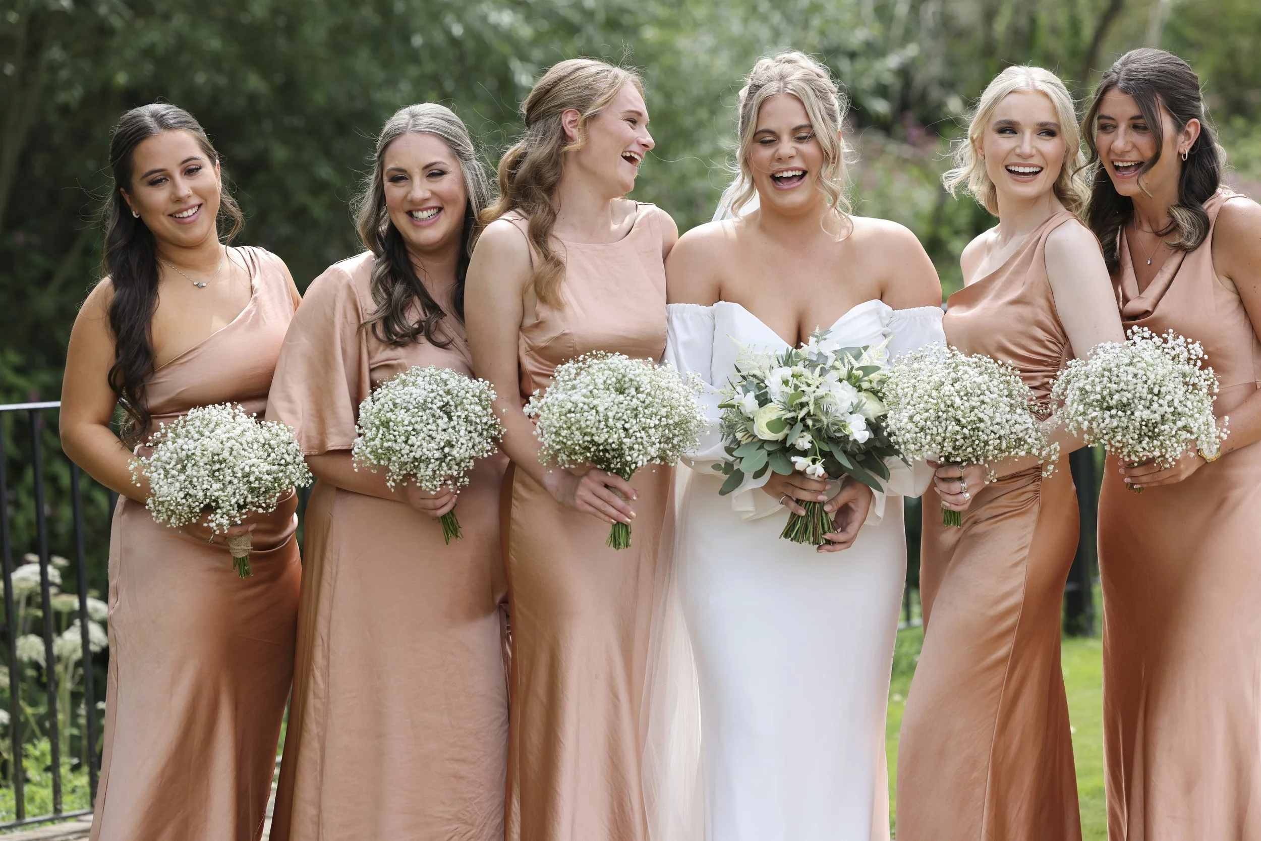 A group of six women, dressed for a wedding, standing outdoors with a lush green background, smiling and laughing with bouquets of white flowers.