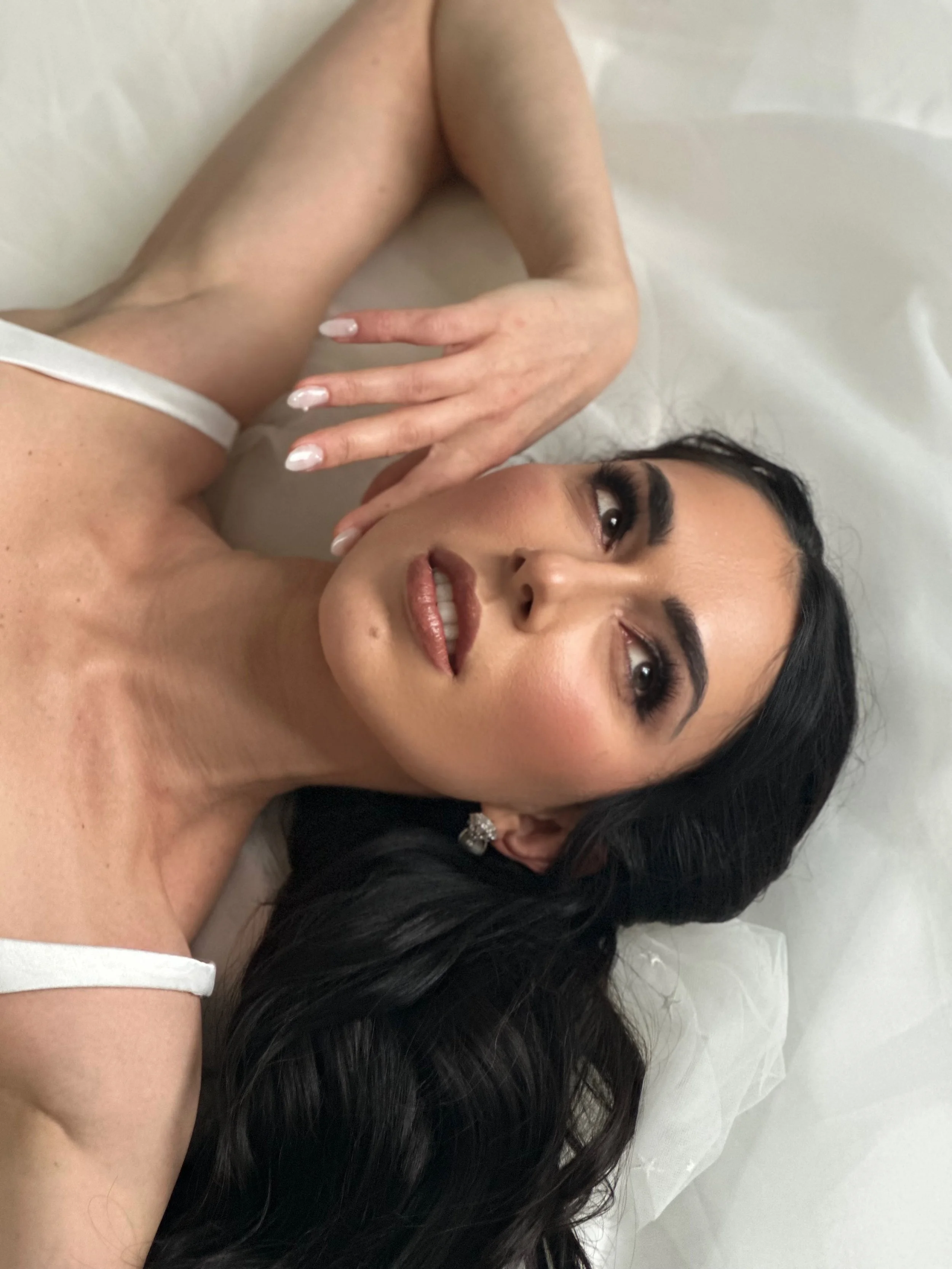 A woman with makeup and dark hair lying on her side on white bedding, wearing a white tank top, with her hand near her face.