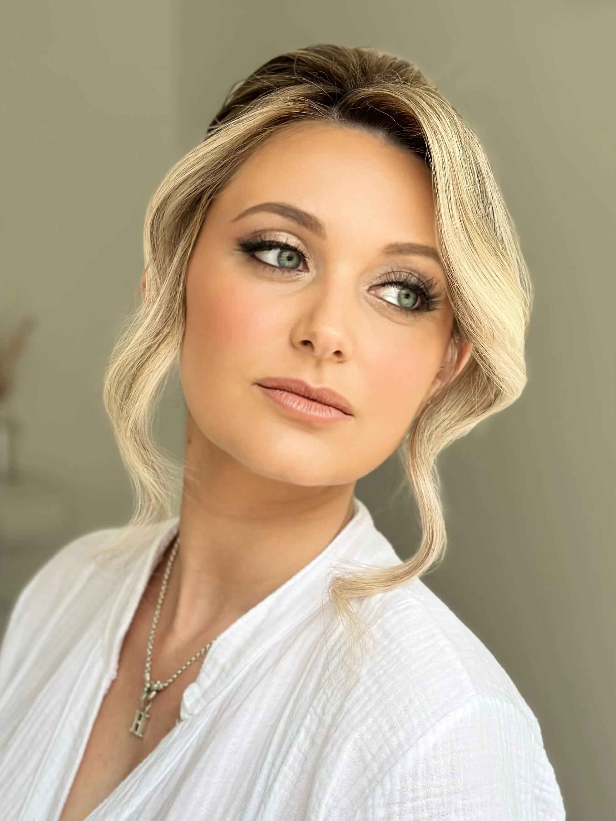 Close-up of a woman with blonde hair styled in loose waves, wearing makeup with defined eyebrows, eyeshadow, and nude lipstick, dressed in a white textured top and a necklace with crosses, standing indoors against a neutral background.