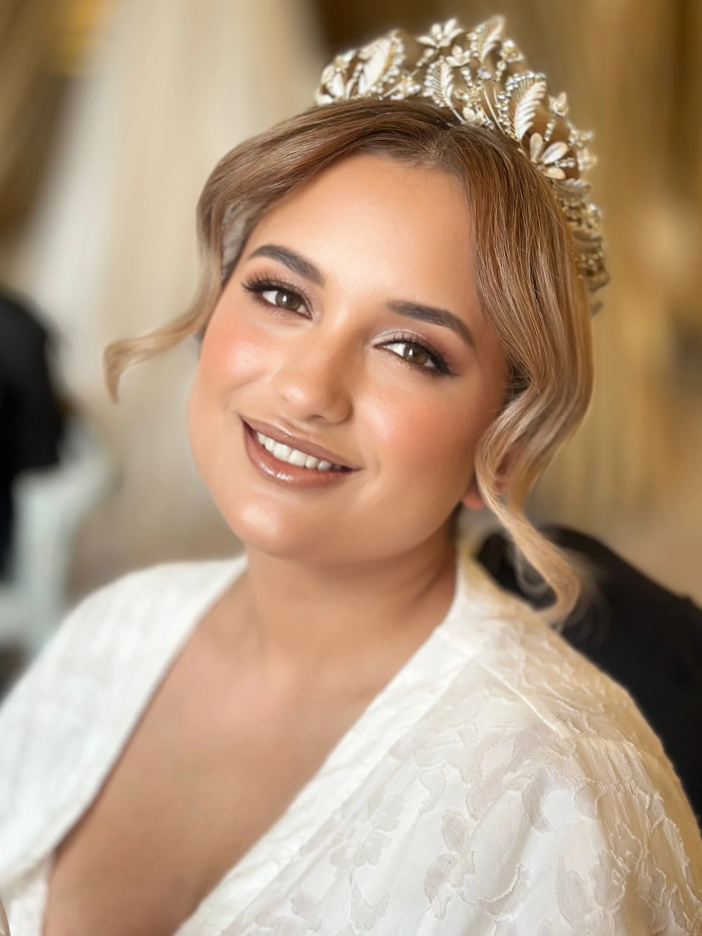 Close-up portrait of a smiling woman with brown hair styled in loose curls, wearing a decorative tiara and a white floral lace dress.