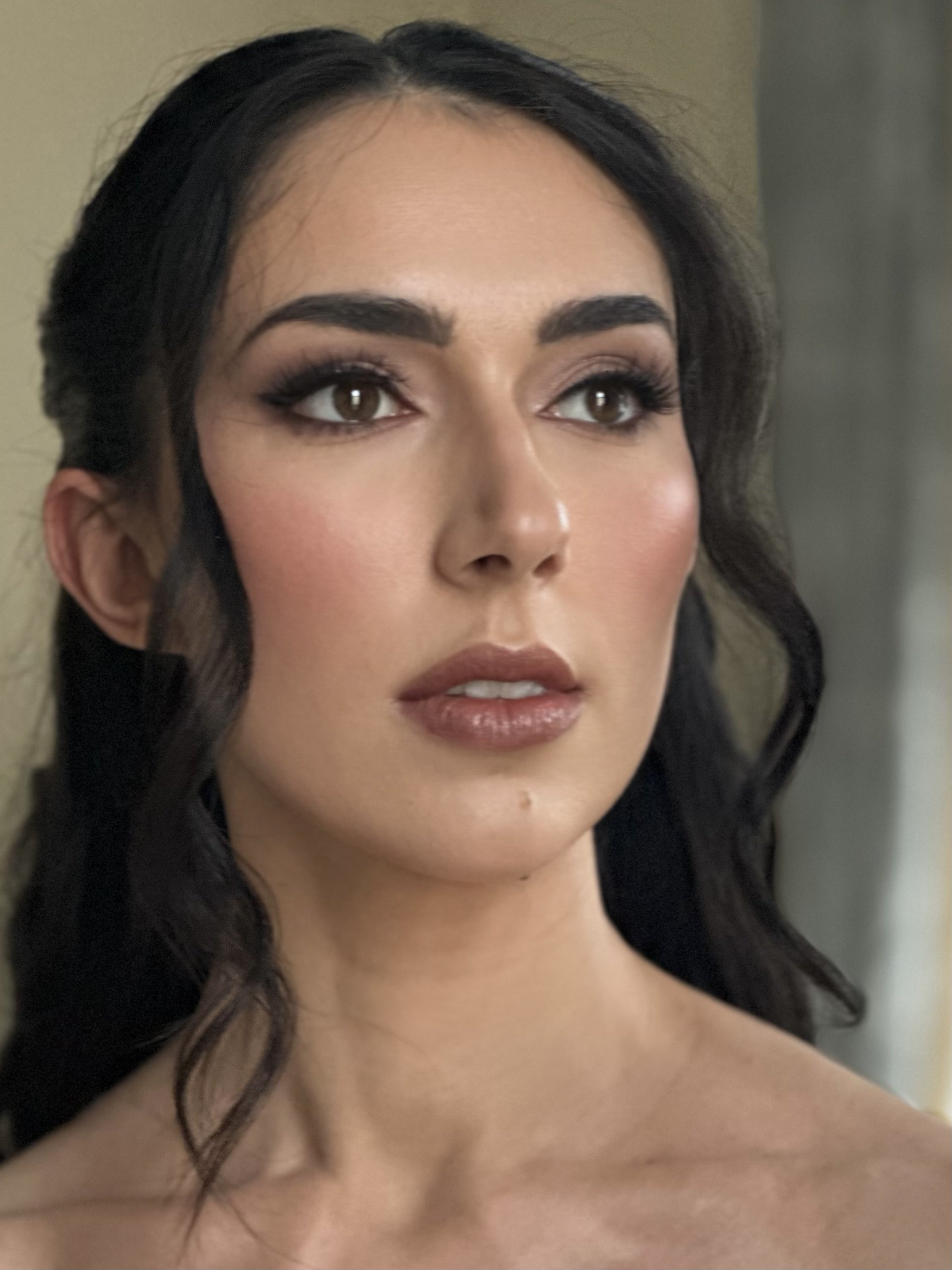 A close-up of a woman with dark brown hair, makeup, and nude lipstick, looking slightly off-camera.