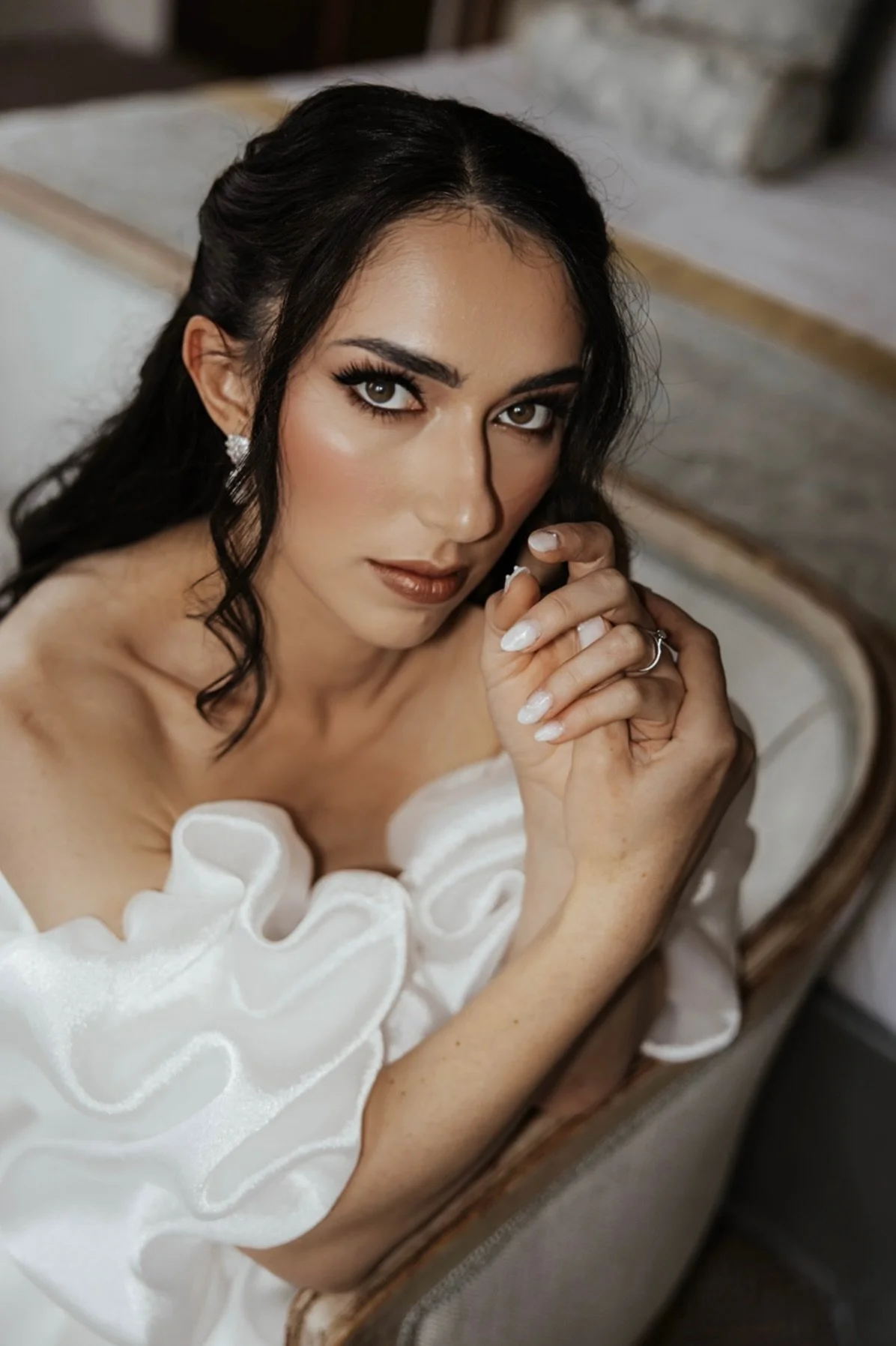 A woman with dark hair, makeup, and jewelry is looking into a mirror, holding her face gently, wearing a white dress with ruffled sleeves, at a vanity or bathroom.