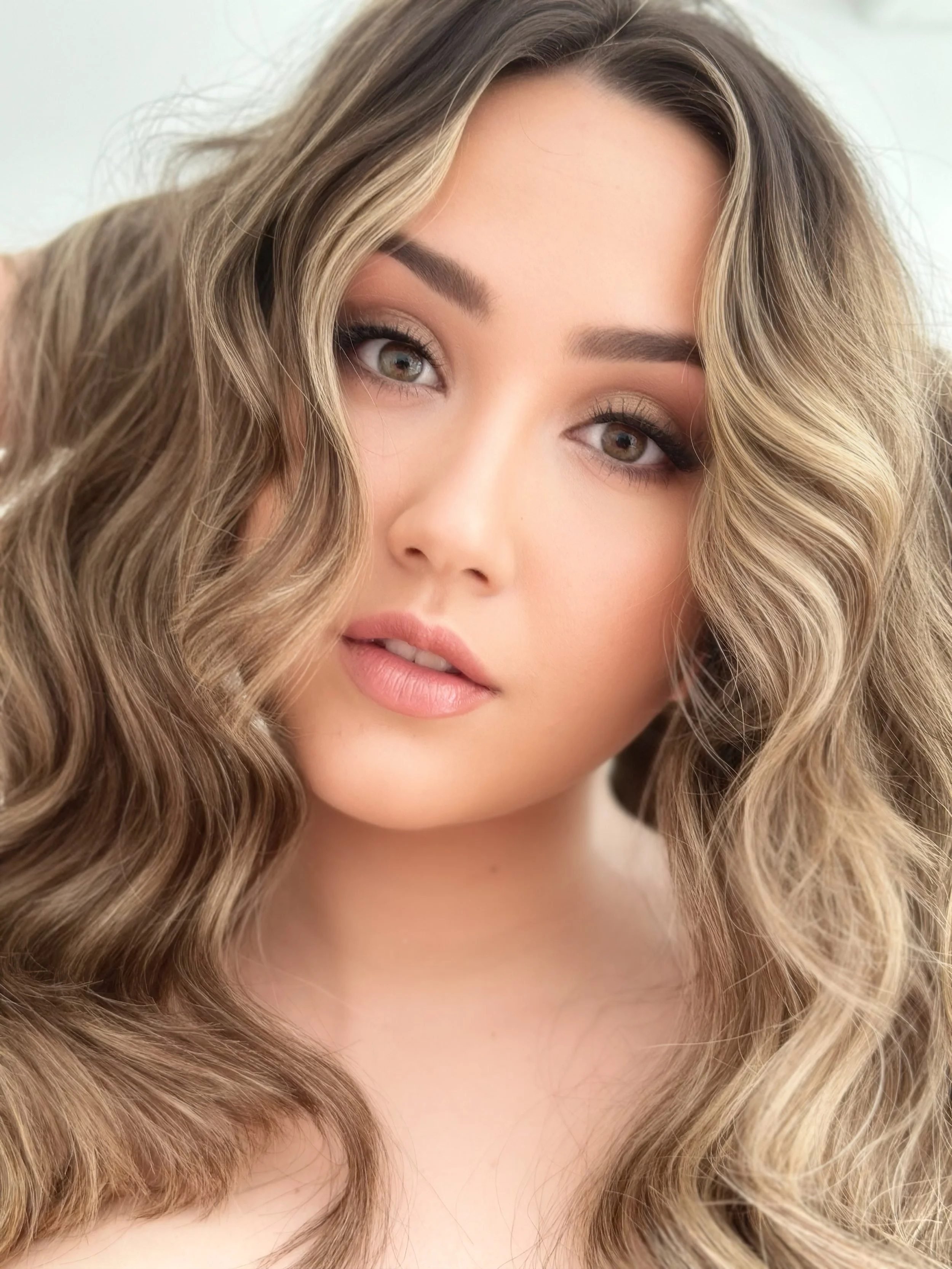 A close-up portrait of a woman with long, wavy blonde hair, hazel eyes, and natural makeup.