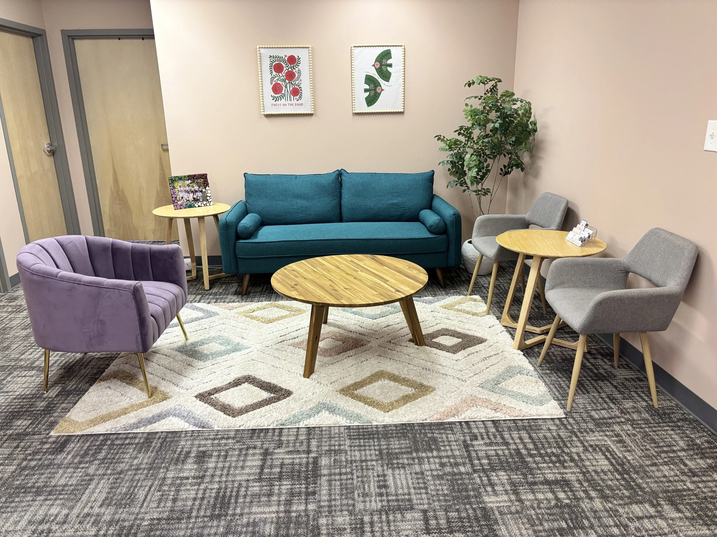 Waiting room with a teal sofa, two beige chairs, a purple armchair, and two round wooden tables on a multicolored patterned rug. There are two framed posters on the wall, a potted plant in the corner, and a family photo on one of the tables.