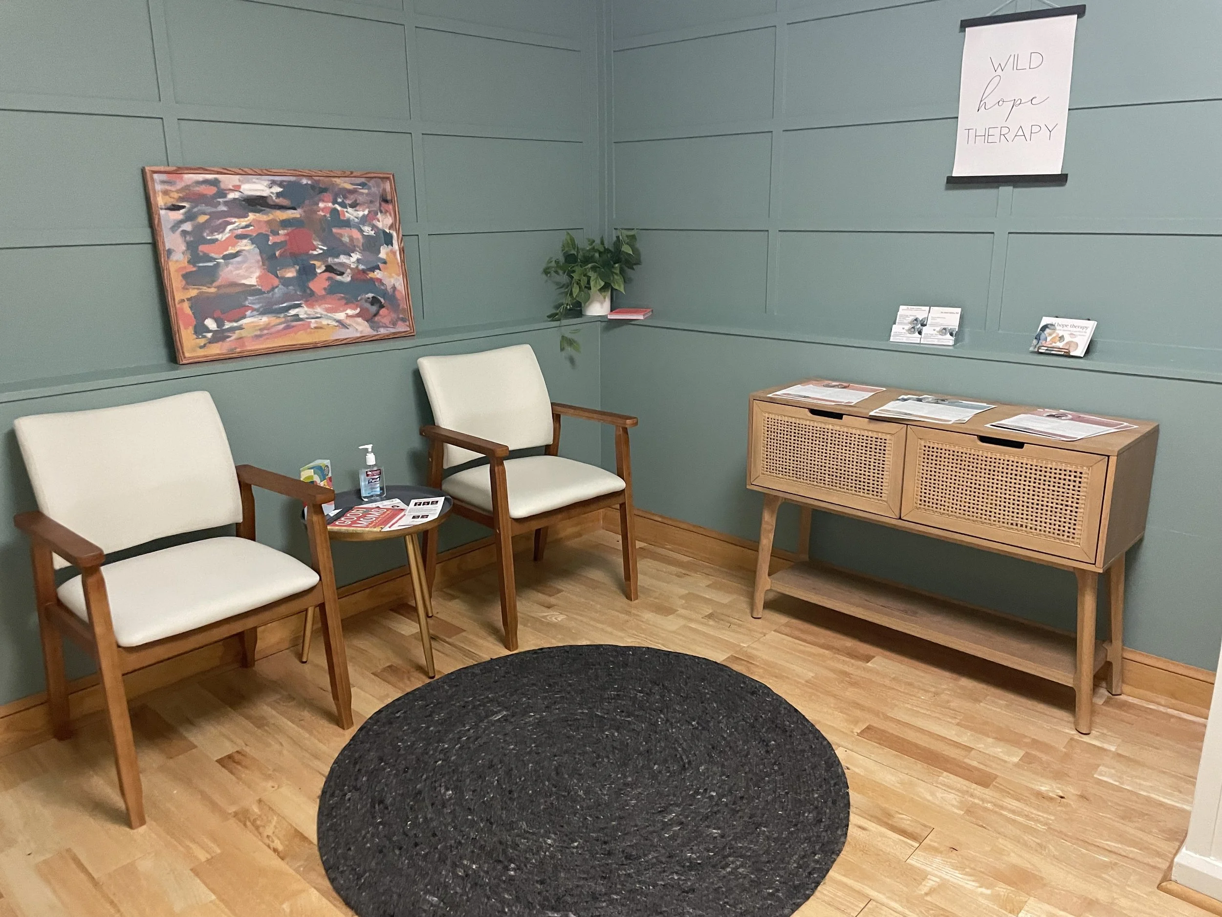 A waiting room with three cream-colored chairs with wooden arms and legs, a small side table holding magazines and hand sanitizer, a green wall with a colorful abstract painting, a potted plant, brochures, and a sign that reads 'WILD hope THERAPY.'