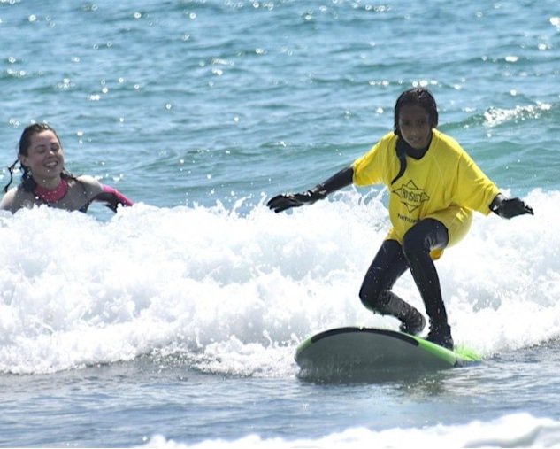 A surfer stands on their board while their instructor cheers them on in the waves behind them