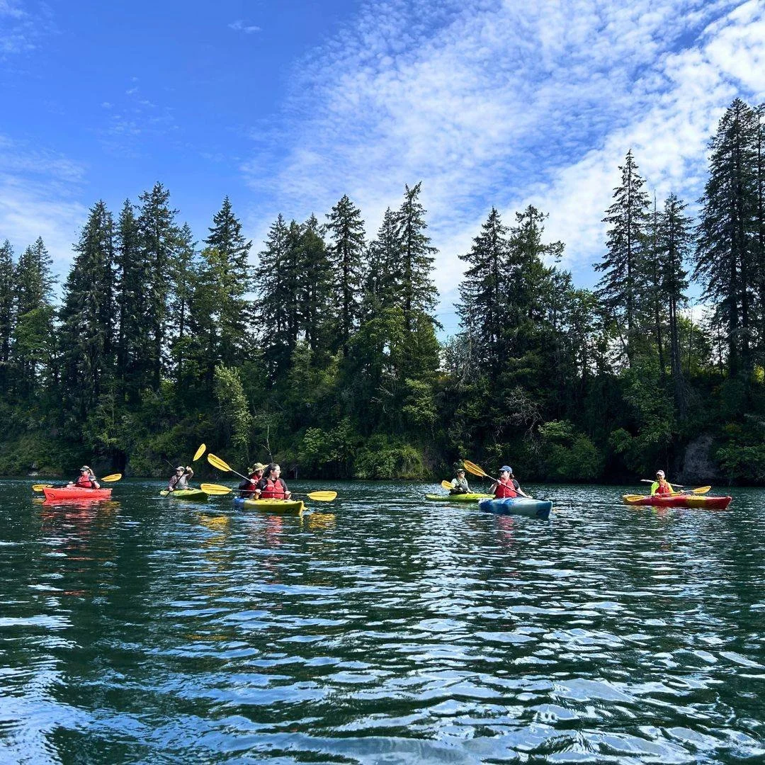 This weekend is our first kayak trip of the season! We're grateful to get out on the water with our community!

Image Description: 7 people in single and tandem (2 person) kayaks paddle on a calm, tree-lined lake on a blue sky day. Everyone is wearin
