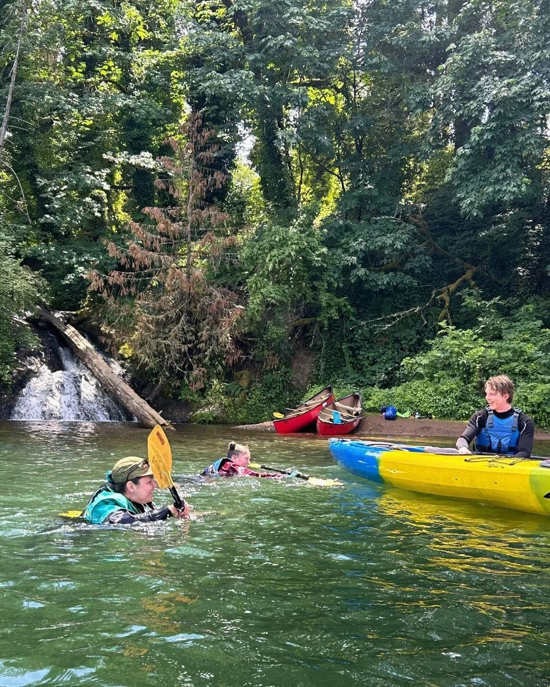 We're one month away from our Staff Training! Summer is just around the bend!

Image Description: Two A-W-L guides wearing P-F-Ds (lifejackets) swim in calm water towards an empty tandem (2 person) kayak while holding paddles. A third guide wearing a