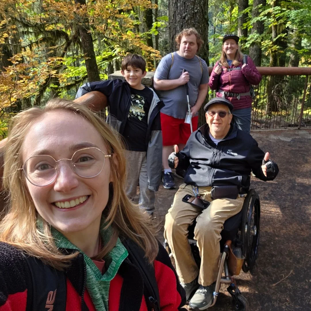 1 person takes a selfie with 4 other people and fall foliage in the background. 1 person is using a wheelchair and 1 person is using a cane.