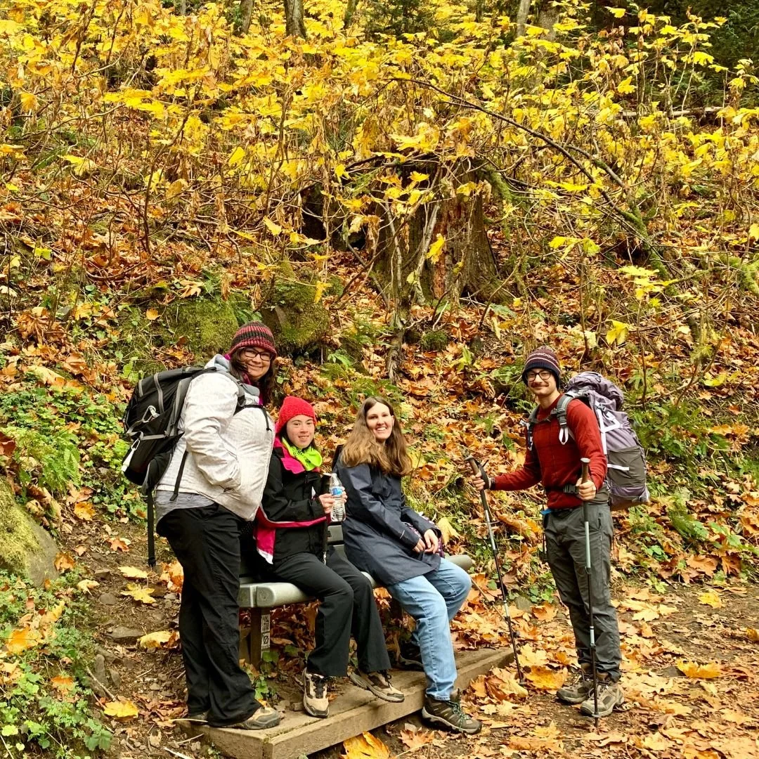 A group of 4 hikers wearing warm layers and backpacks pose together on a trail with colorful fall foliage.