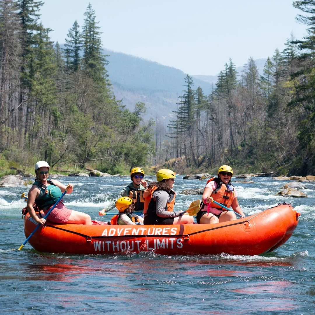 5 people in a brightly colored raft wearing helmets and P-F-Ds (lifejackets) smile and paddle on a sunny day