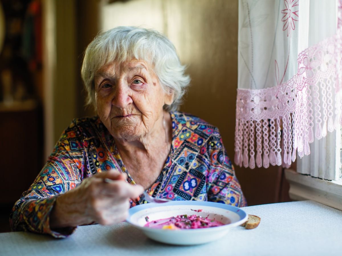 Una anciana se sienta a la mesa a tomar sopa. Lleva una camisa estampada de colores y se detiene a mirar al fotógrafo.