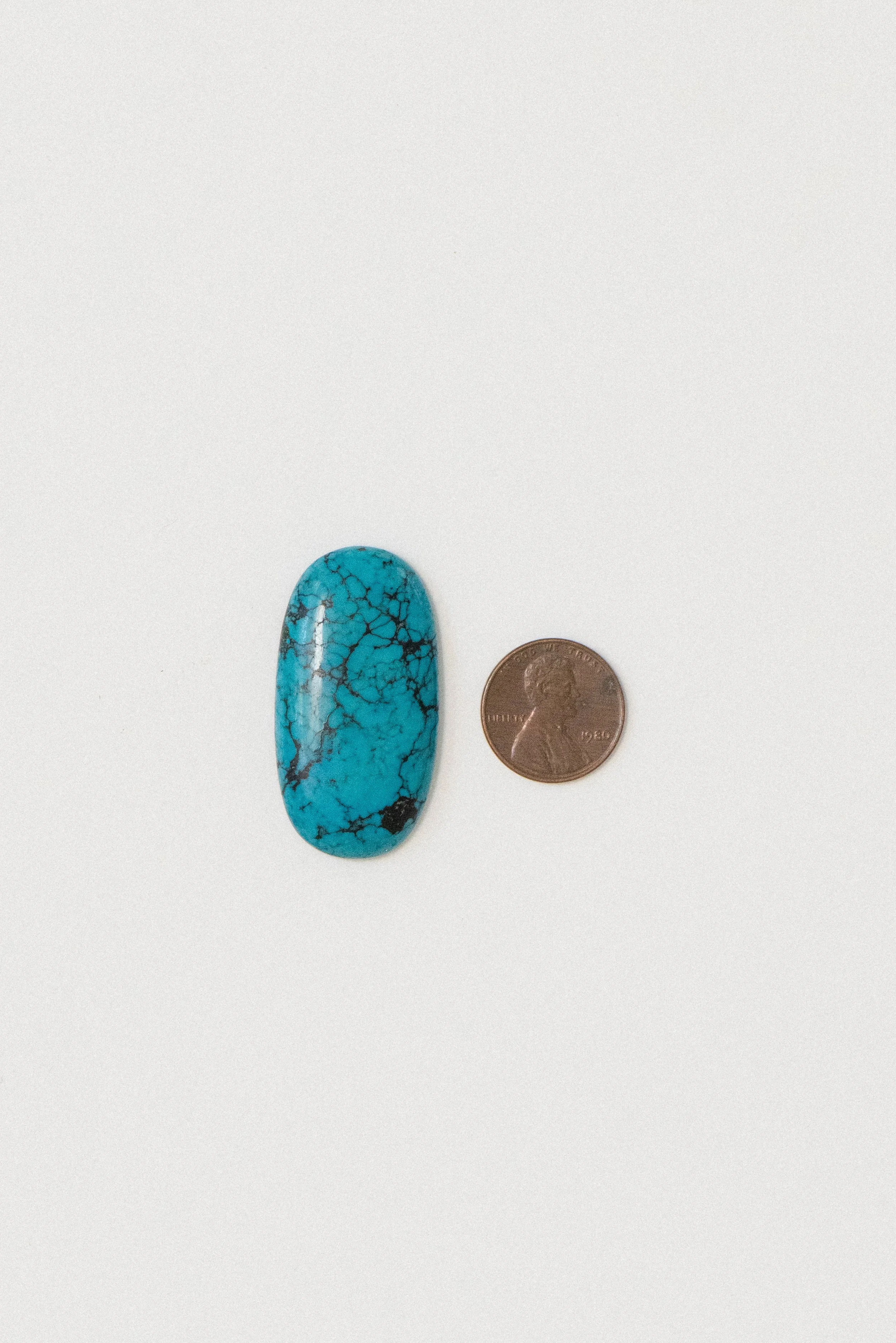 Polished turquoise cabochon from Sinai, Egypt with black matrix, shown next to a penny for size reference on a white background.