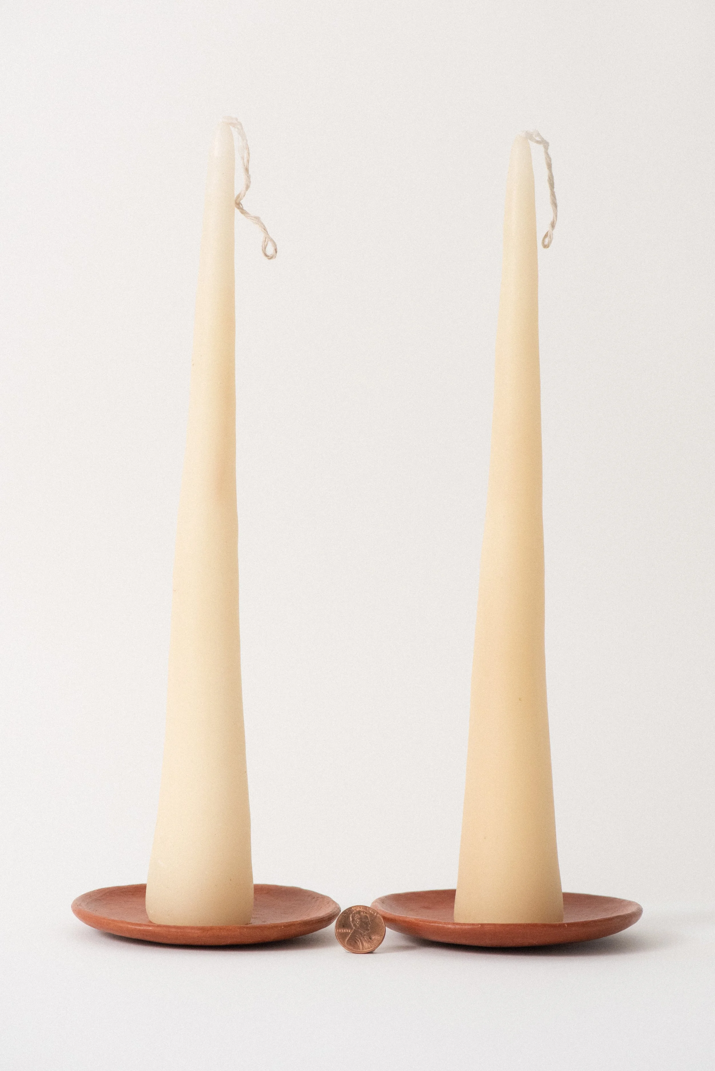 Oaxaca beeswax taper candles on red clay barro rojo plates shown with a penny for scale on a white background.