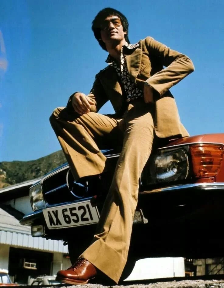 Bruce Lee wearing a tan flared suit and floral shirt, sitting confidently on a vintage red Mercedes with license plate AX 6521, under a clear blue sky in the 1970s