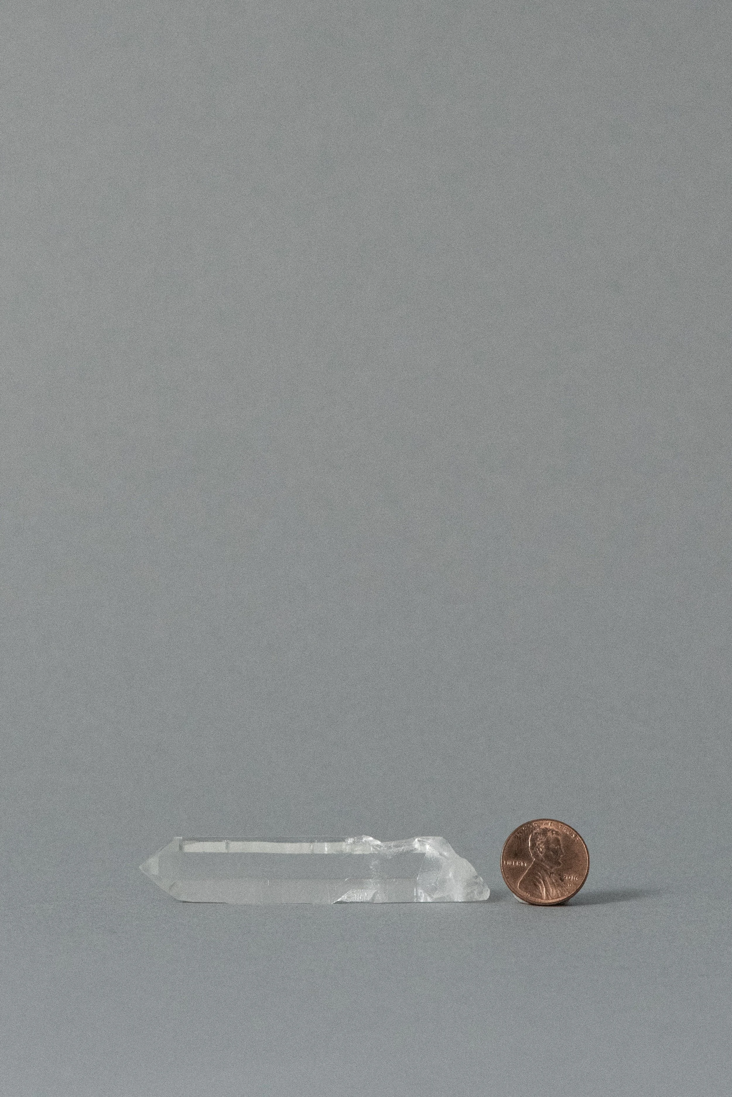 Single-terminated clear quartz point from Brazil shown next to a penny for scale, photographed on a gray background.