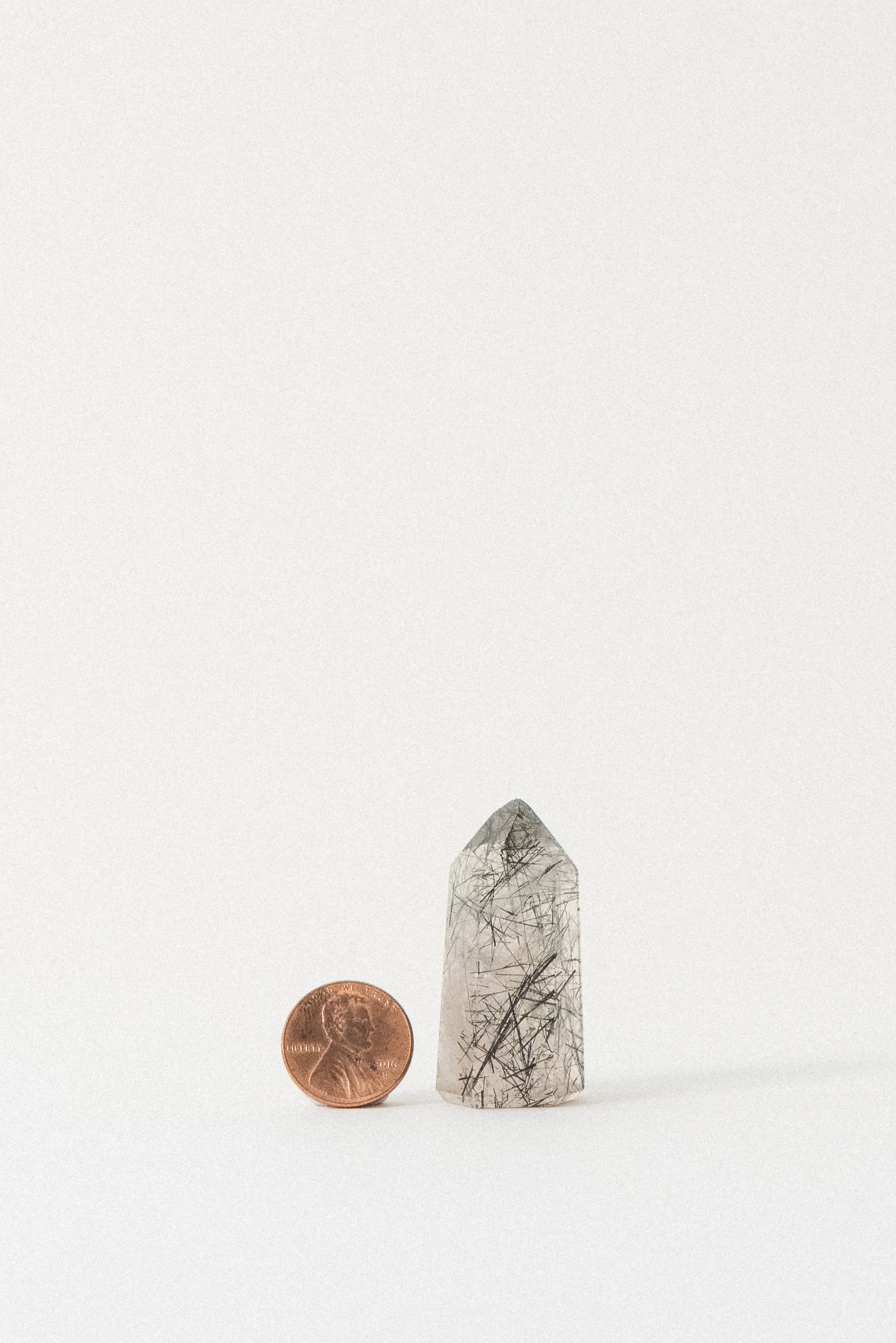 Black tourmaline included quartz tower with needle-like inclusions, shown upright beside a penny for scale. Offered by Crystal Voyager.