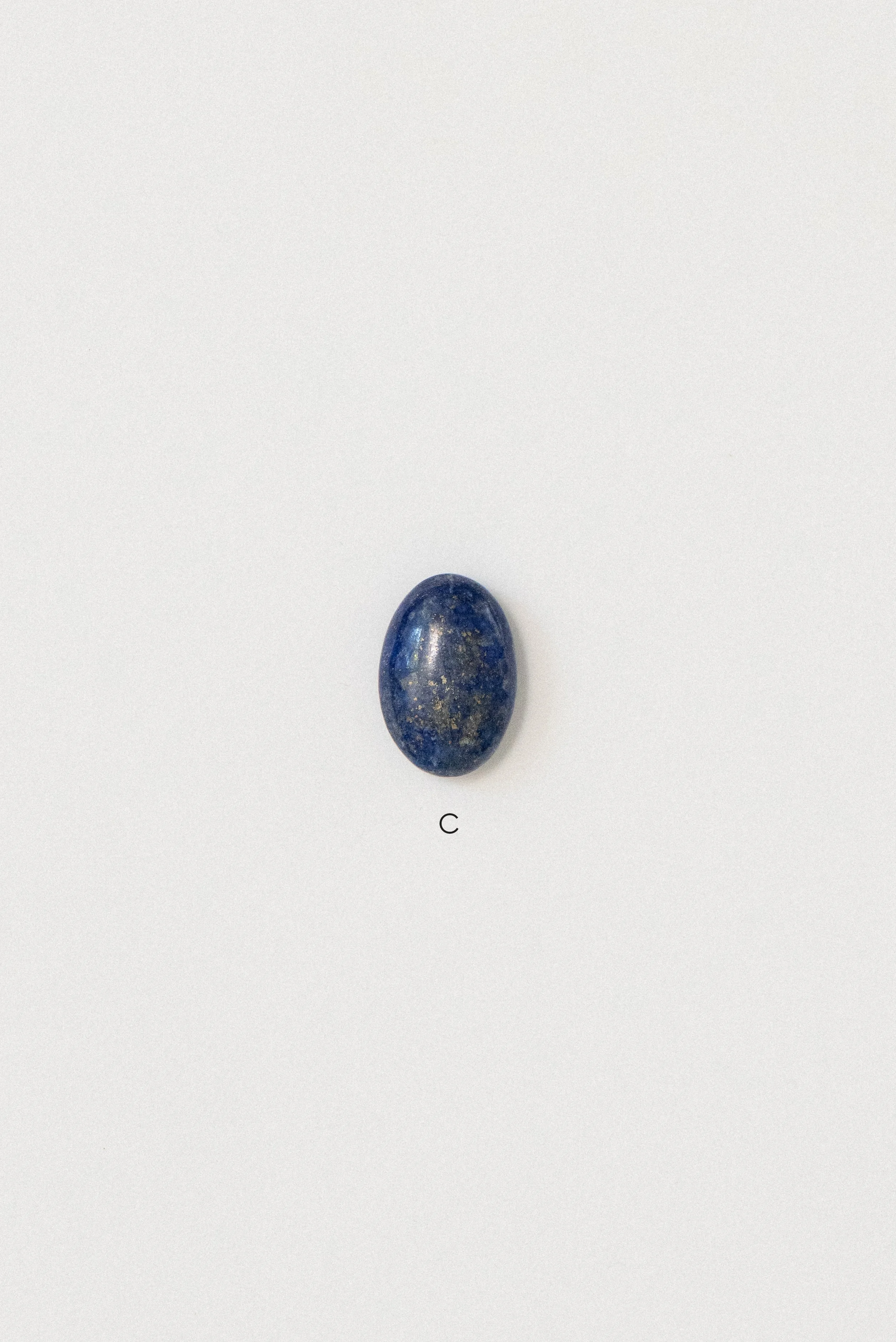 Single polished lapis lazuli stone from Afghanistan labeled 'C' on a white background, featuring an oval shape with visible golden pyrite inclusions.