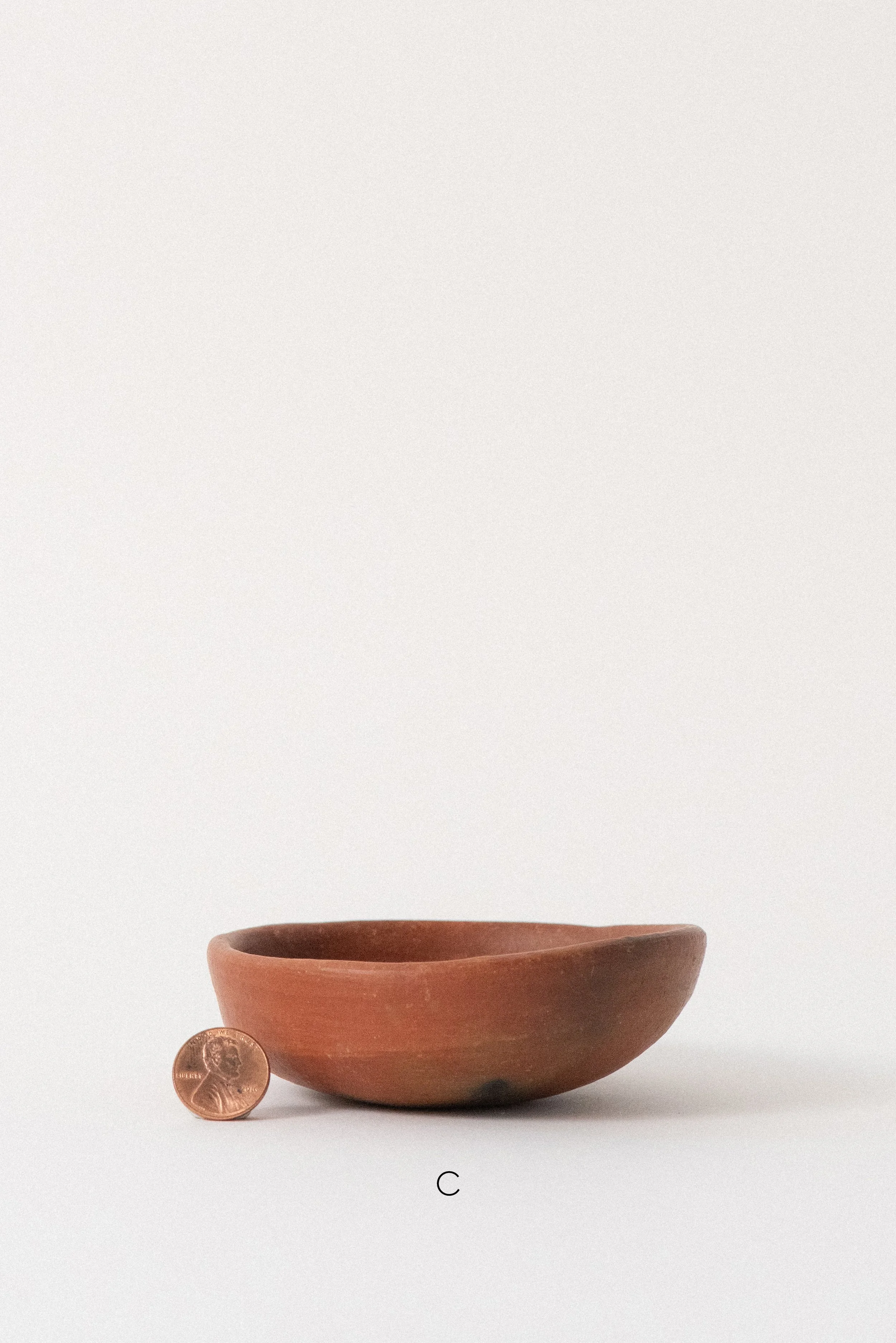 Single handmade barro rojo red clay bowl labeled C, crafted in Oaxaca, Mexico, shown with a penny for scale against a white background — rustic and versatile kitchenware.