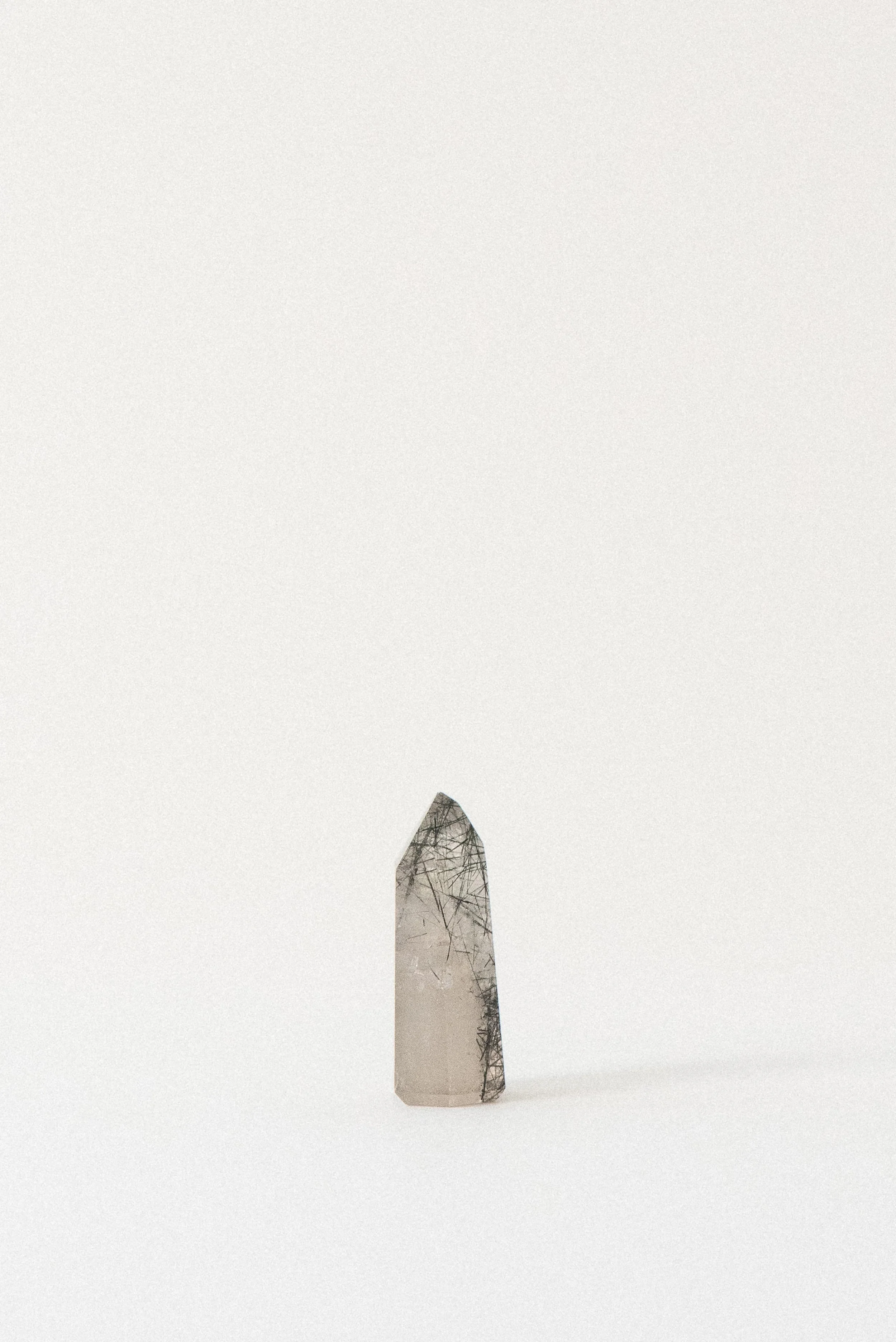 Side view of a clear quartz tower with needle-like black tourmaline inclusions, photographed on a soft white background. Origin: Brazil.
