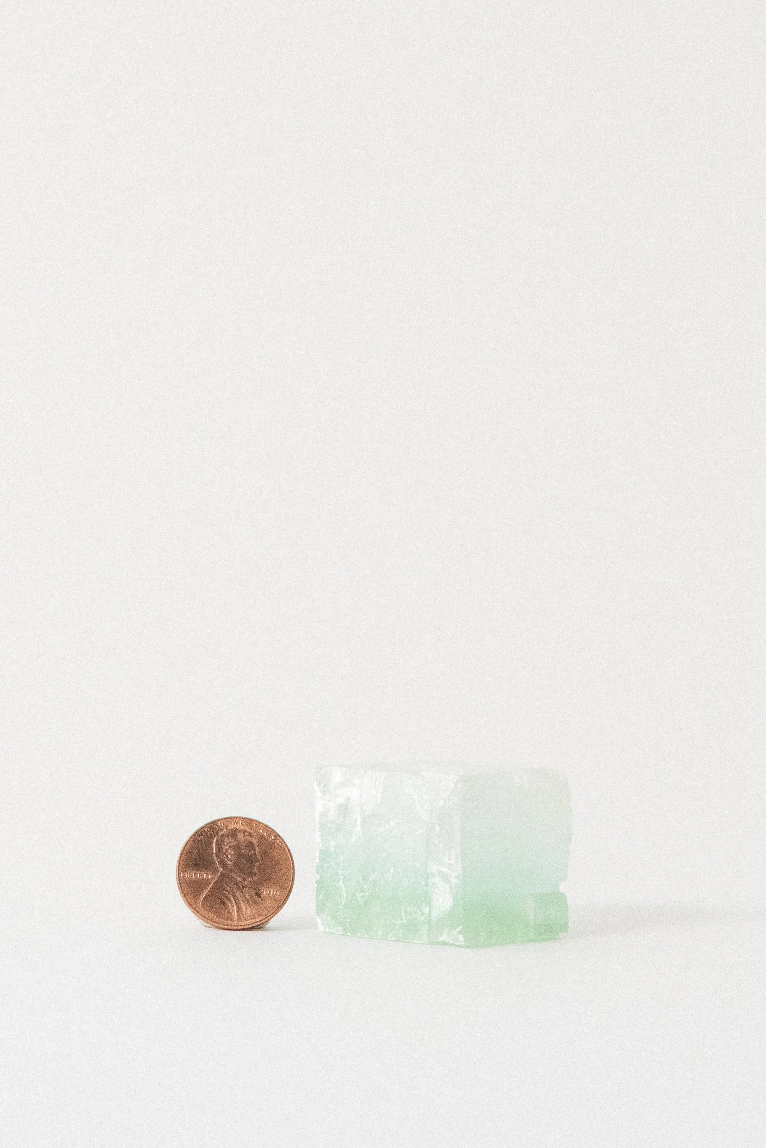 Small green calcite rhomboid crystal from Mexico shown next to a penny for scale, displayed on a soft white background with subtle light gradients