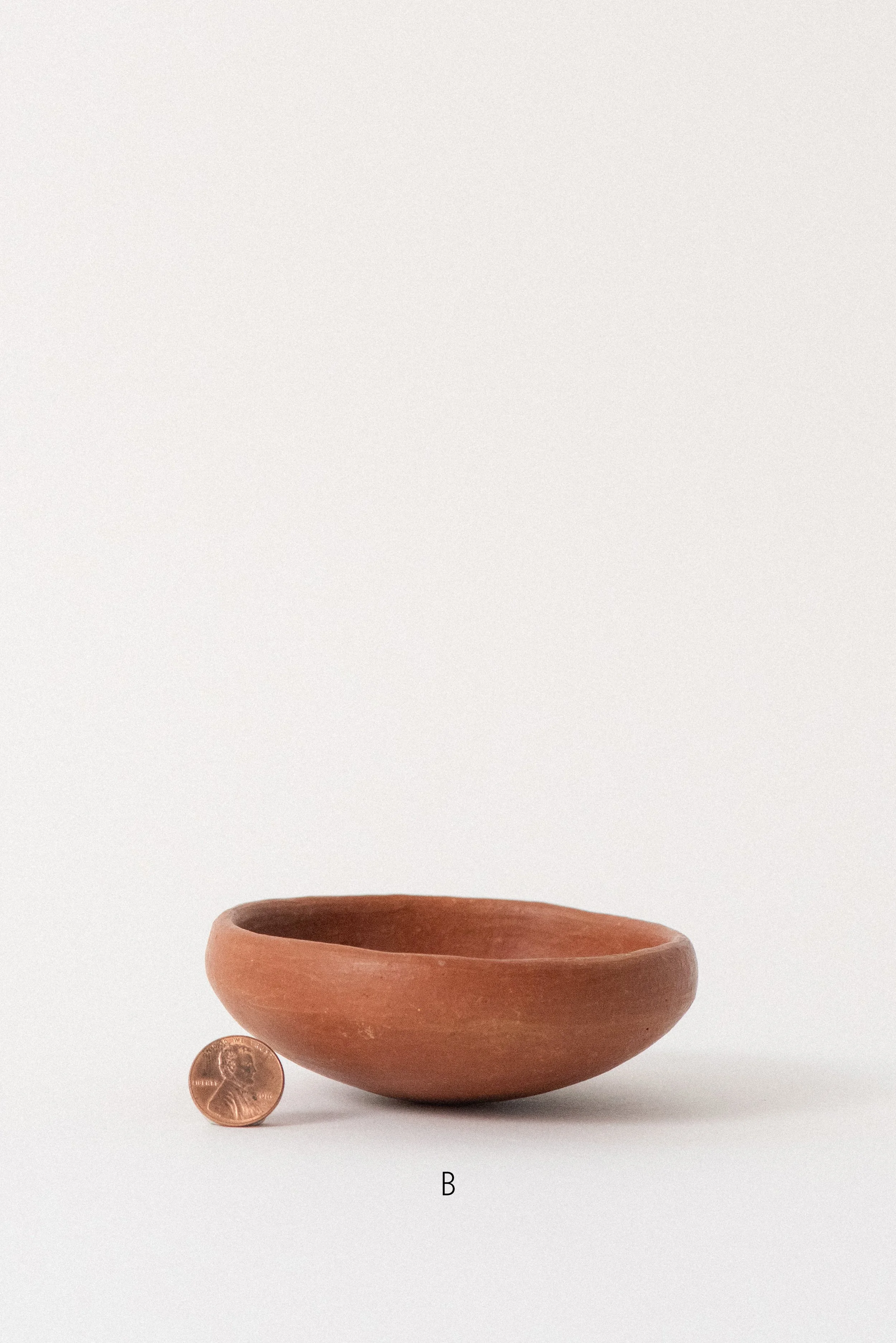 Single handmade barro rojo red clay bowl labeled B, crafted in Oaxaca, Mexico, shown with a penny for scale against a white background — rustic and versatile kitchenware.
