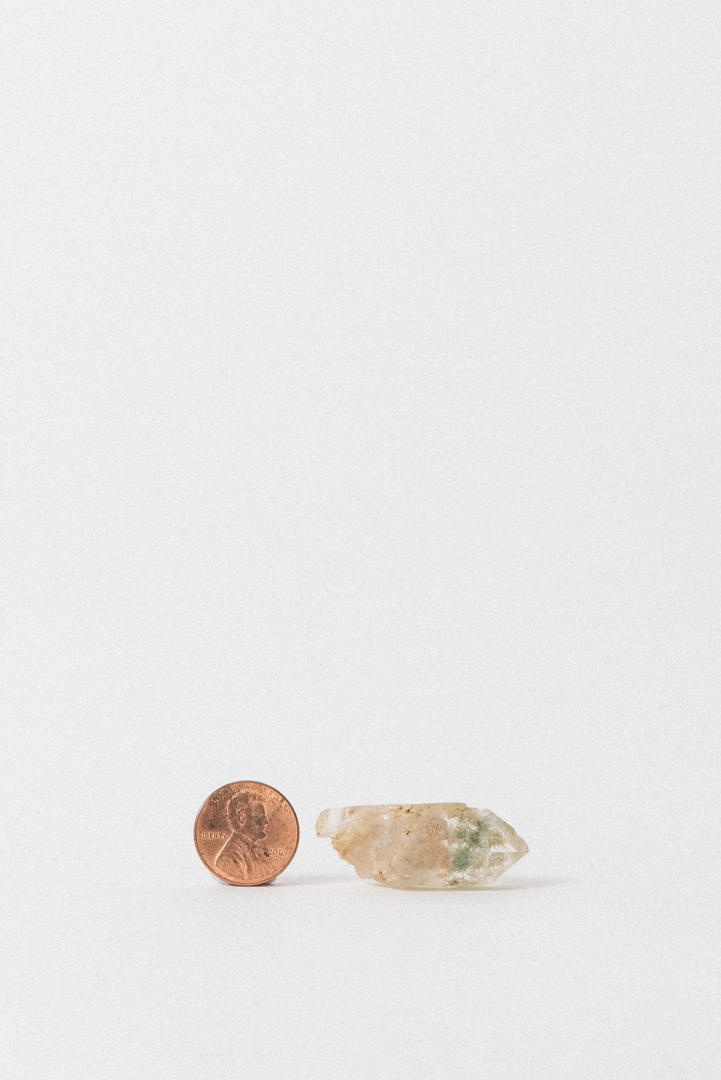 Clear quartz point with green pumpellyite inclusions next to a penny for scale, showing its small size. Photographed on a white background.