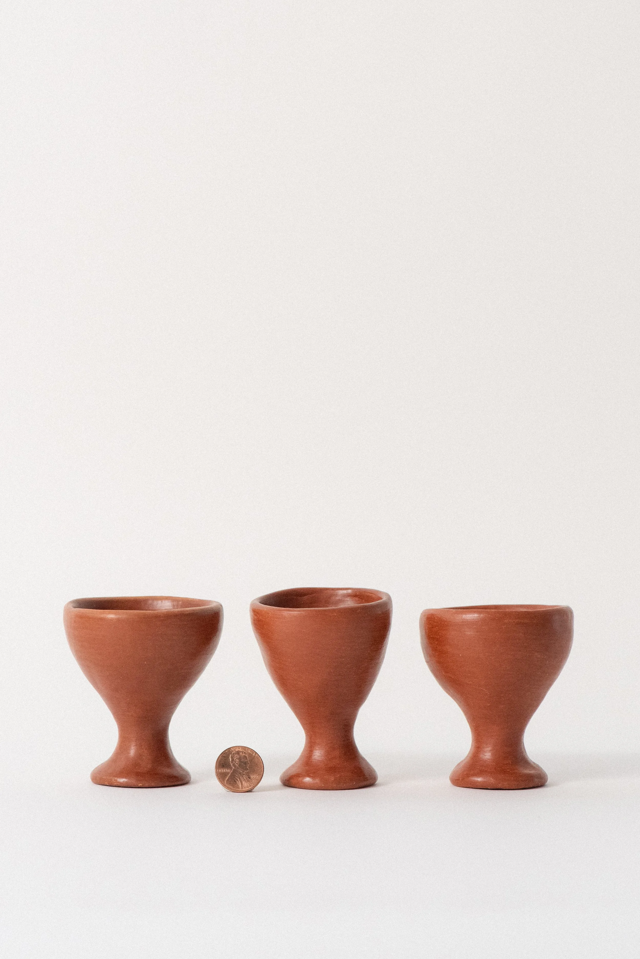 Set of three handmade barro rojo red clay egg cups from Oaxaca, Mexico, shown with a penny for scale against a white background — artisanal and functional tableware.