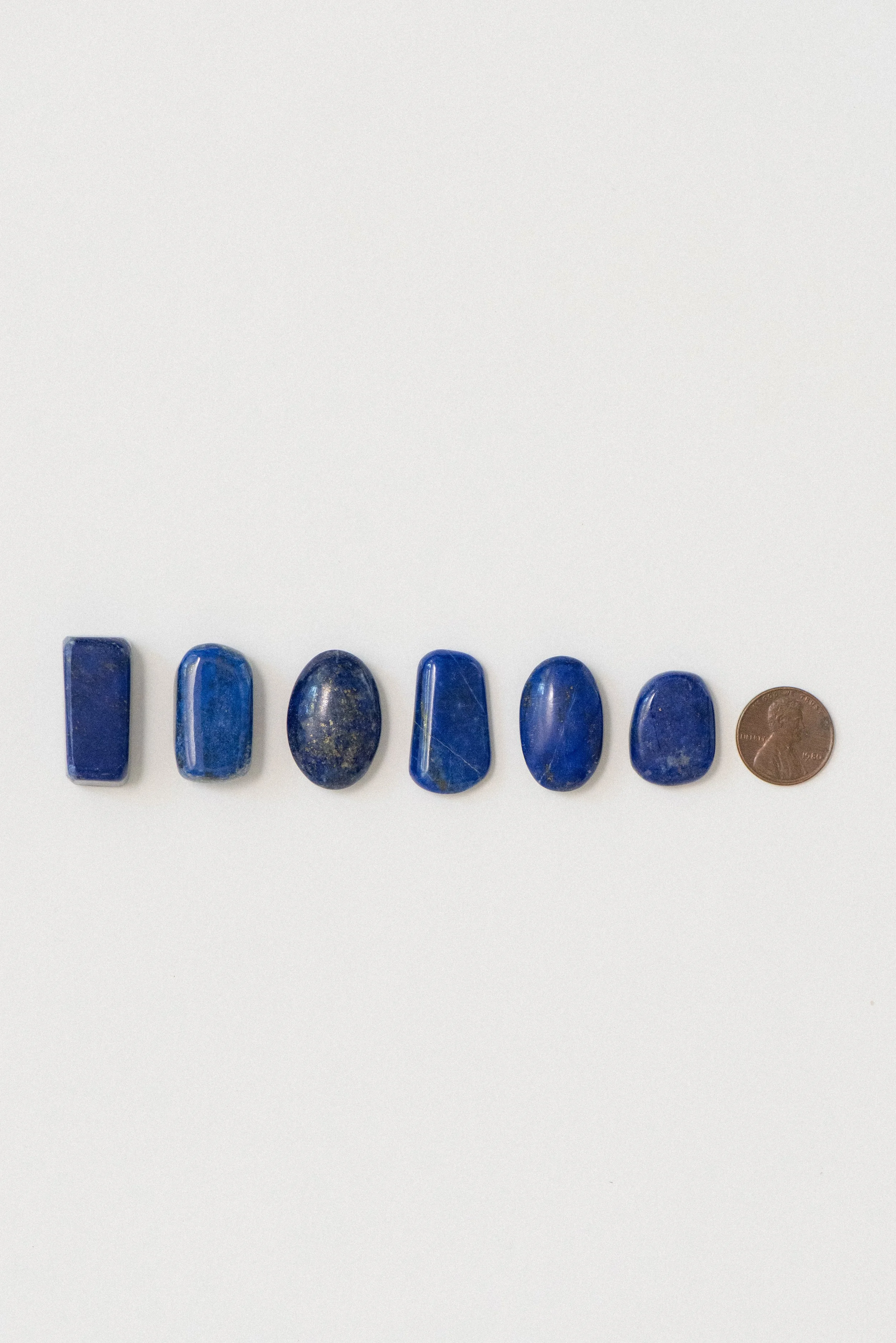 Six natural lapis lazuli polished stones from Afghanistan displayed on a white background with a penny for size reference.