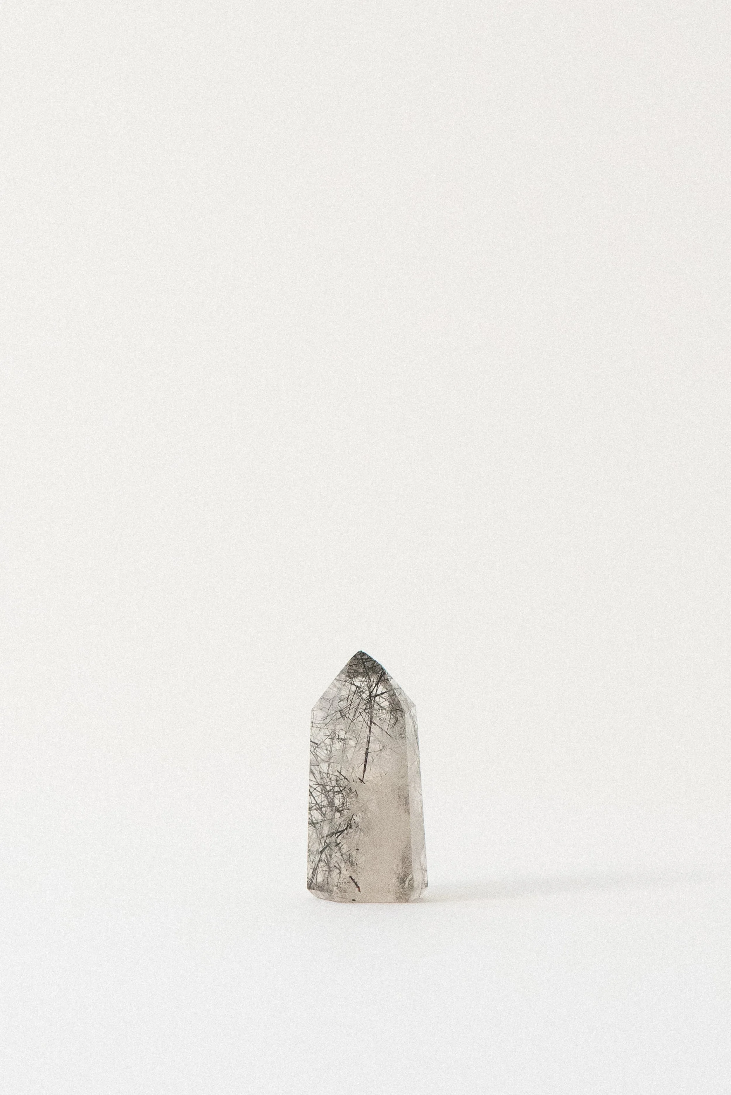 Front view of a polished clear quartz tower with delicate black tourmaline inclusions, photographed on a soft white background. Mined in Brazil.