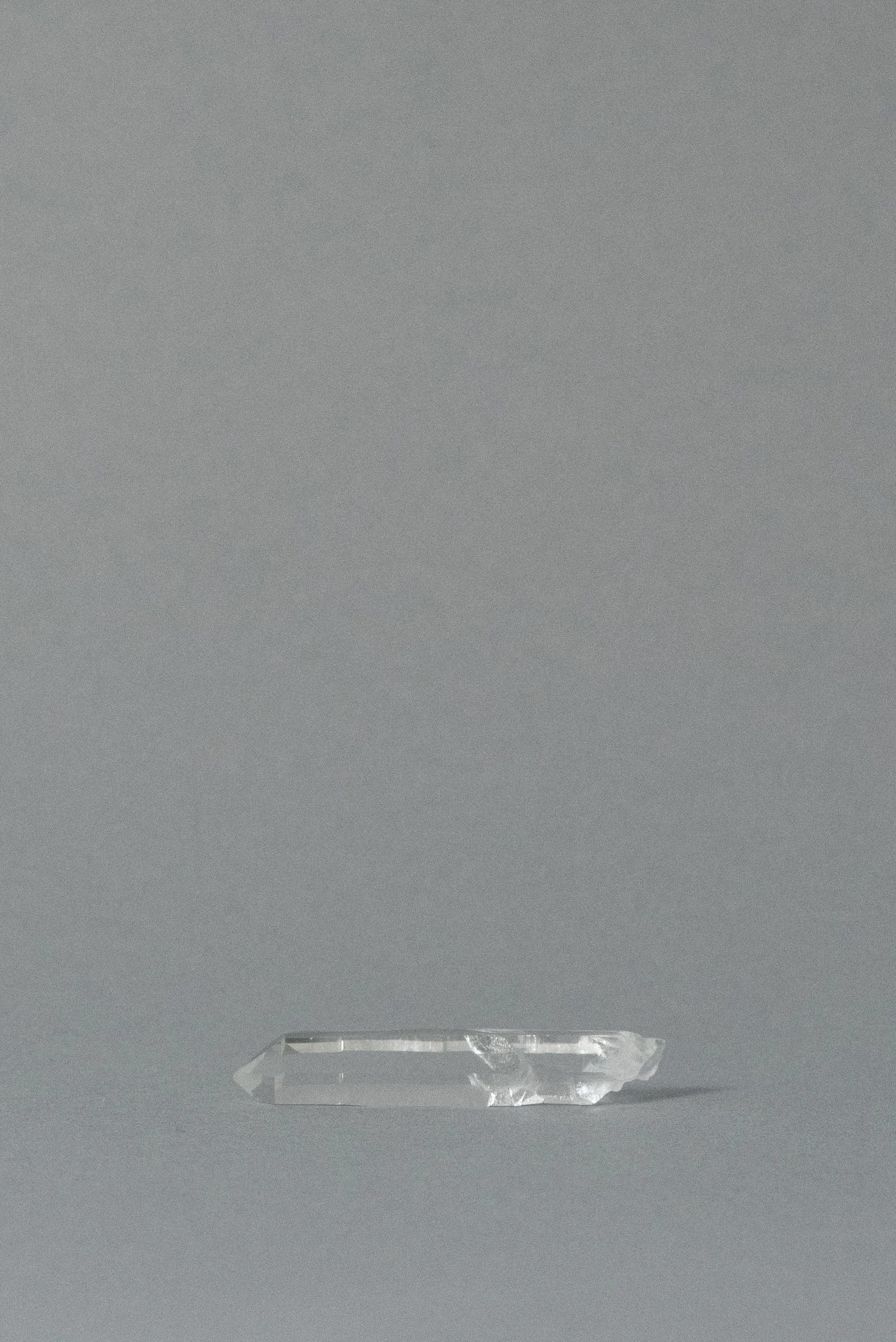 Natural clear quartz point crystal from Brazil, single-terminated and transparent, laid horizontally on a soft gray background.